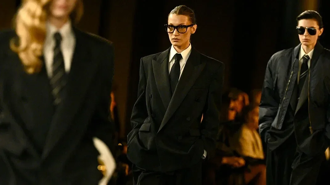 Three women in black suits and ties standing with hands in pockets, wearing sunglasses and serious expressions, in a dimly lit room.