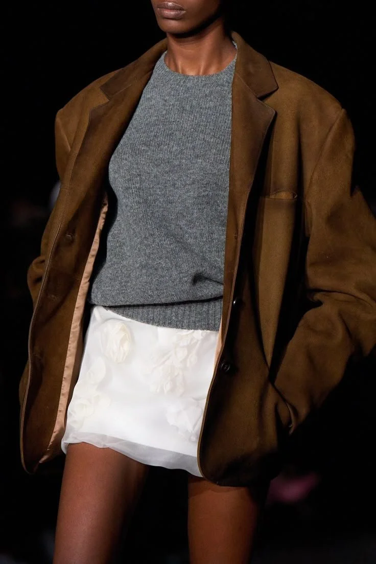 A woman wearing a brown oversized blazer over a grey sweater, with white shorts featuring textured floral patterns.