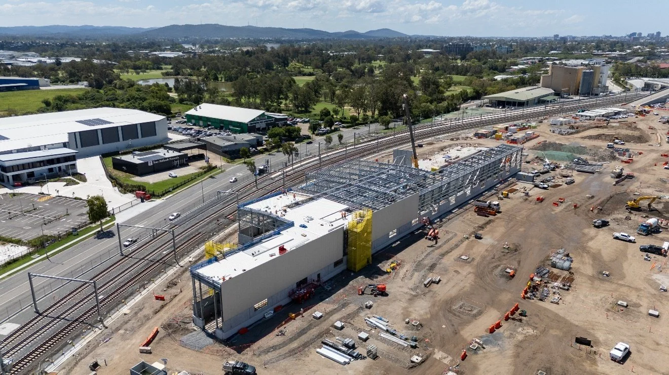 Construction site next to a train track with a partially built modern building, construction equipment, and cars in the surrounding area.