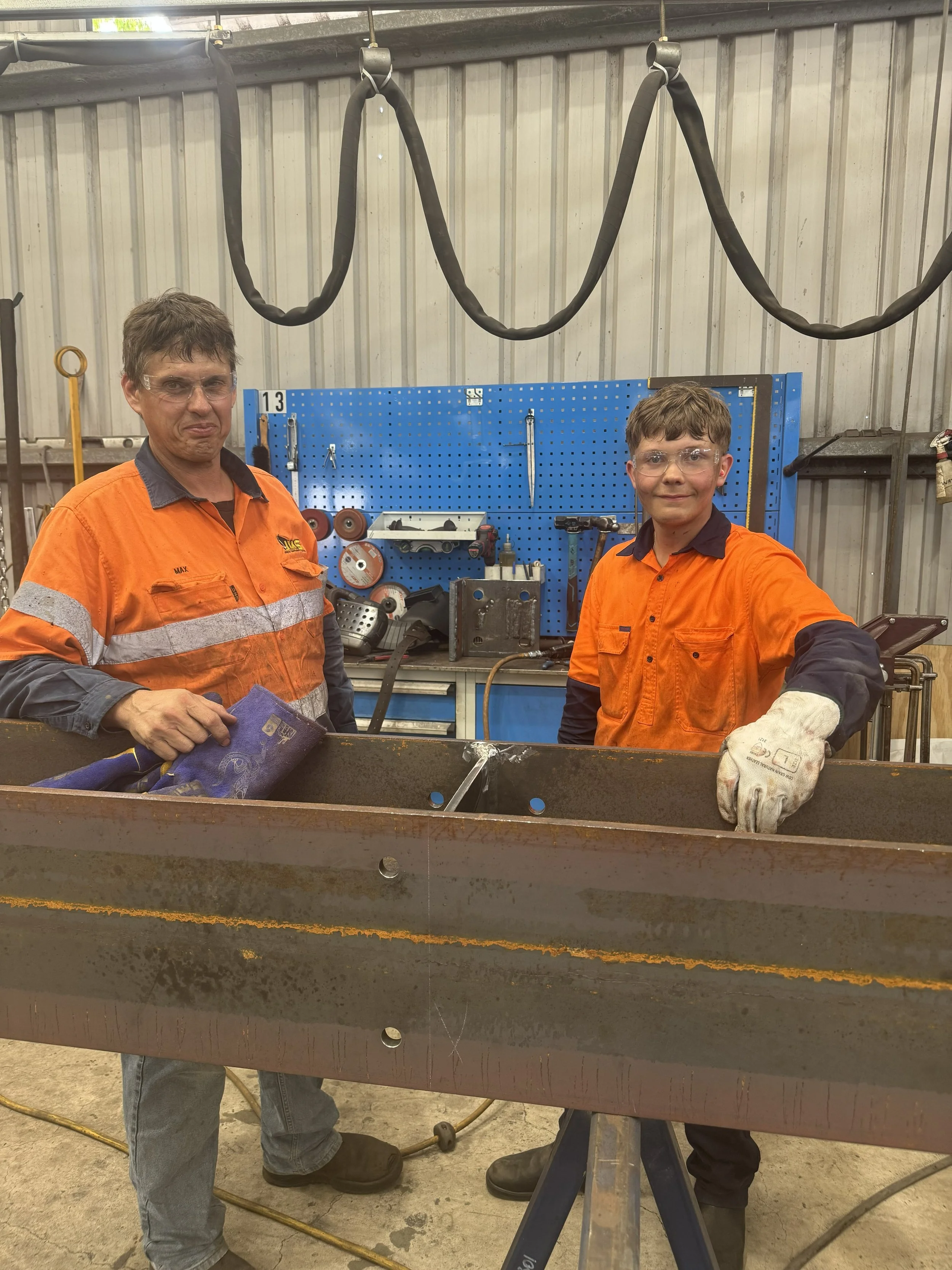 Two workers wearing orange safety shirts and gloves are welding or working on a large metal beam in a workshop. One worker is holding a tool, and a blue pegboard with tools is visible in the background.