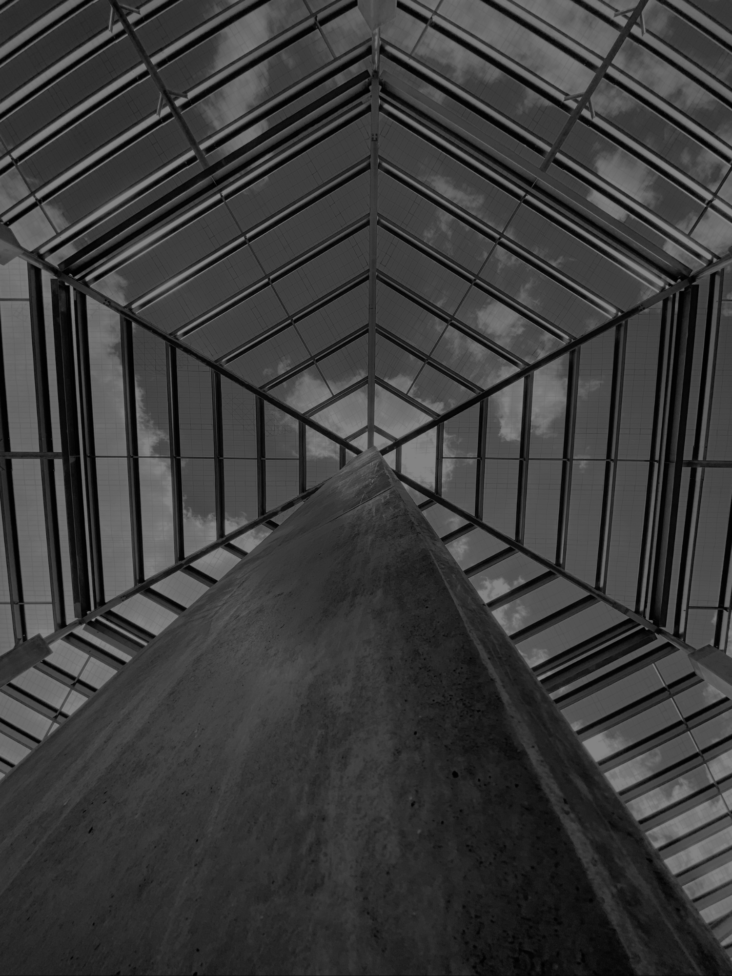 Looking up at the corner of a modern glass roof structure supported by a tall concrete pillar, with clouds visible through the glass panels.