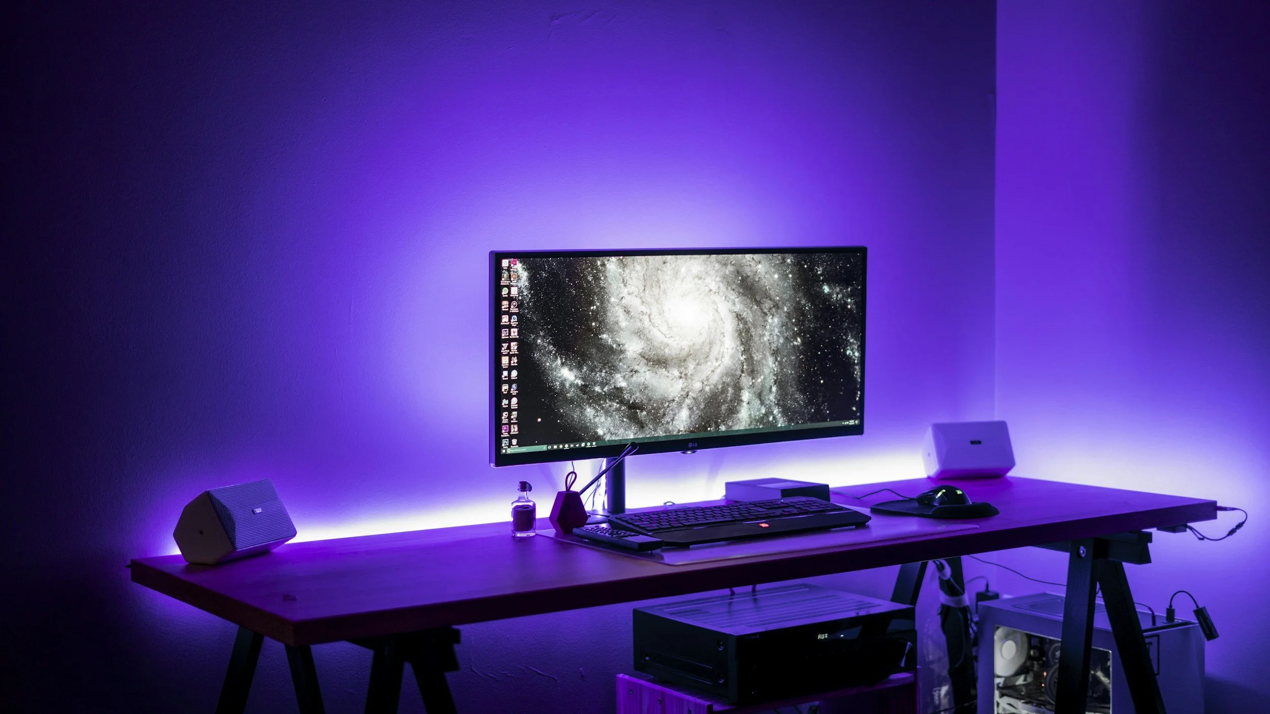 A computer desk setup with a widescreen monitor displaying a galaxy, purple LED ambient lighting behind the desk, and computer accessories like speakers, keyboard, mouse, and a PC case.