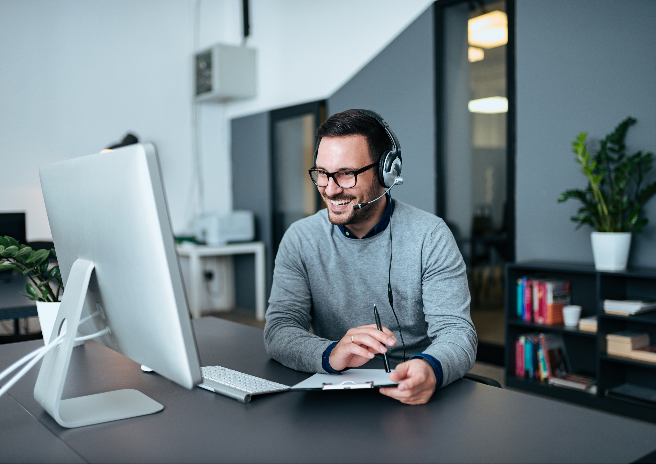 A man in glasses wearing a gray sweater and a headset, smiling at a computer monitor in a modern office space, holding a pen and a notepad.