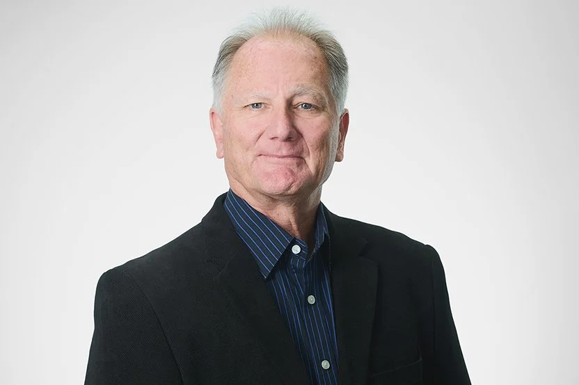 Technical Director, Craig Byrnes, wearing a black blazer and a blue striped shirt, facing the camera with a slight smile against a plain white background.