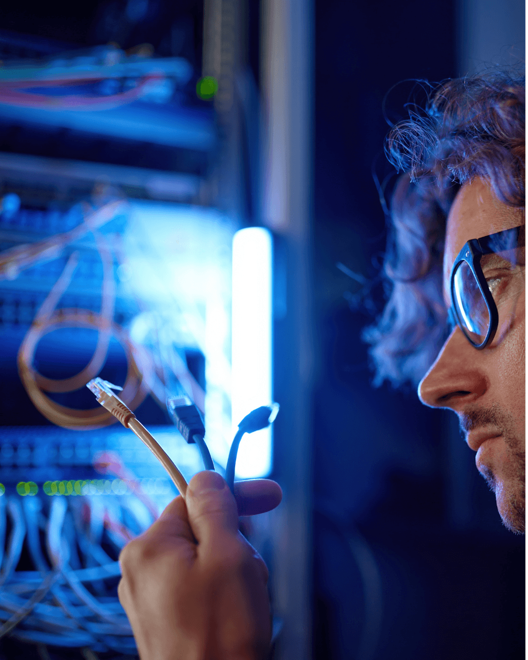 A technician in black glasses handles an Ethernet cable in a server room filled with network equipment and glowing lights.