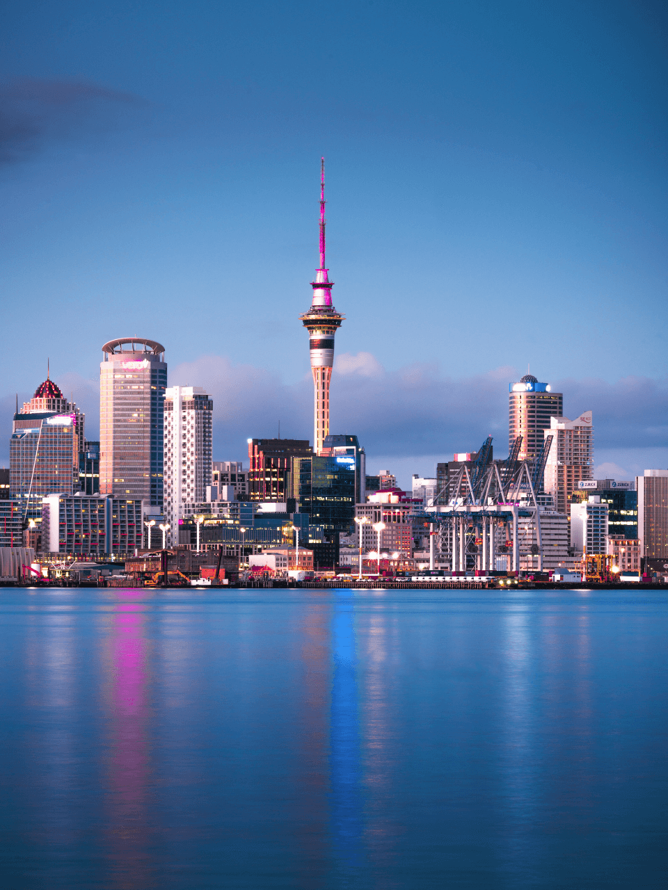 Skyline of downtown Auckland, New Zealand, with the Sky Tower, various high-rise buildings, and an industrial port in the foreground, reflected in the water.