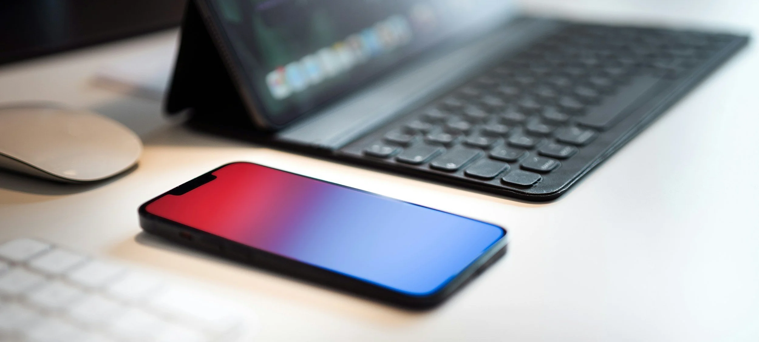 Smartphone with gradient screen resting on a white desk near a computer mouse and a keyboard, with a tablet with keyboard in the background.