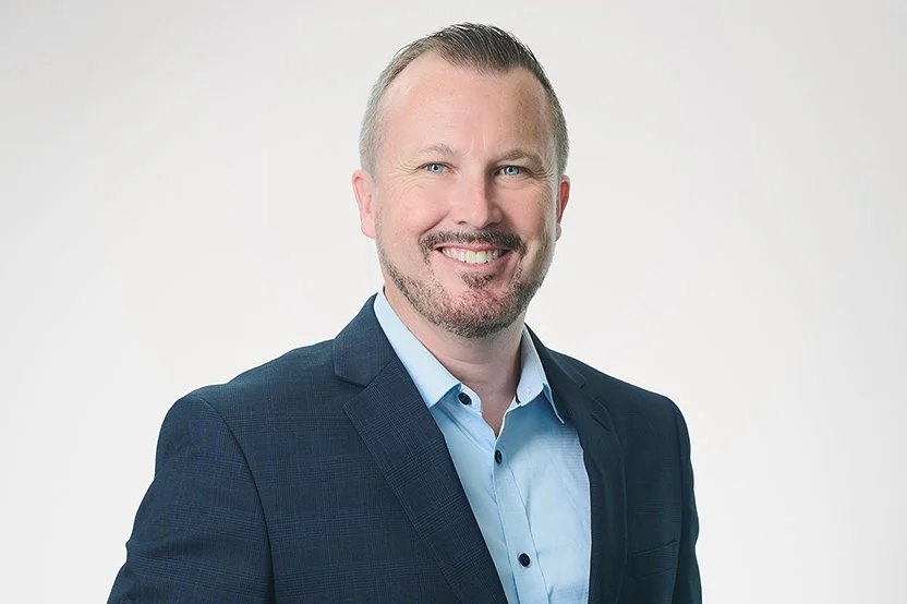 Headshot of a smiling Chris Lee with short light hair and facial hair, wearing a navy blazer and blue collared shirt, standing against a plain white background.