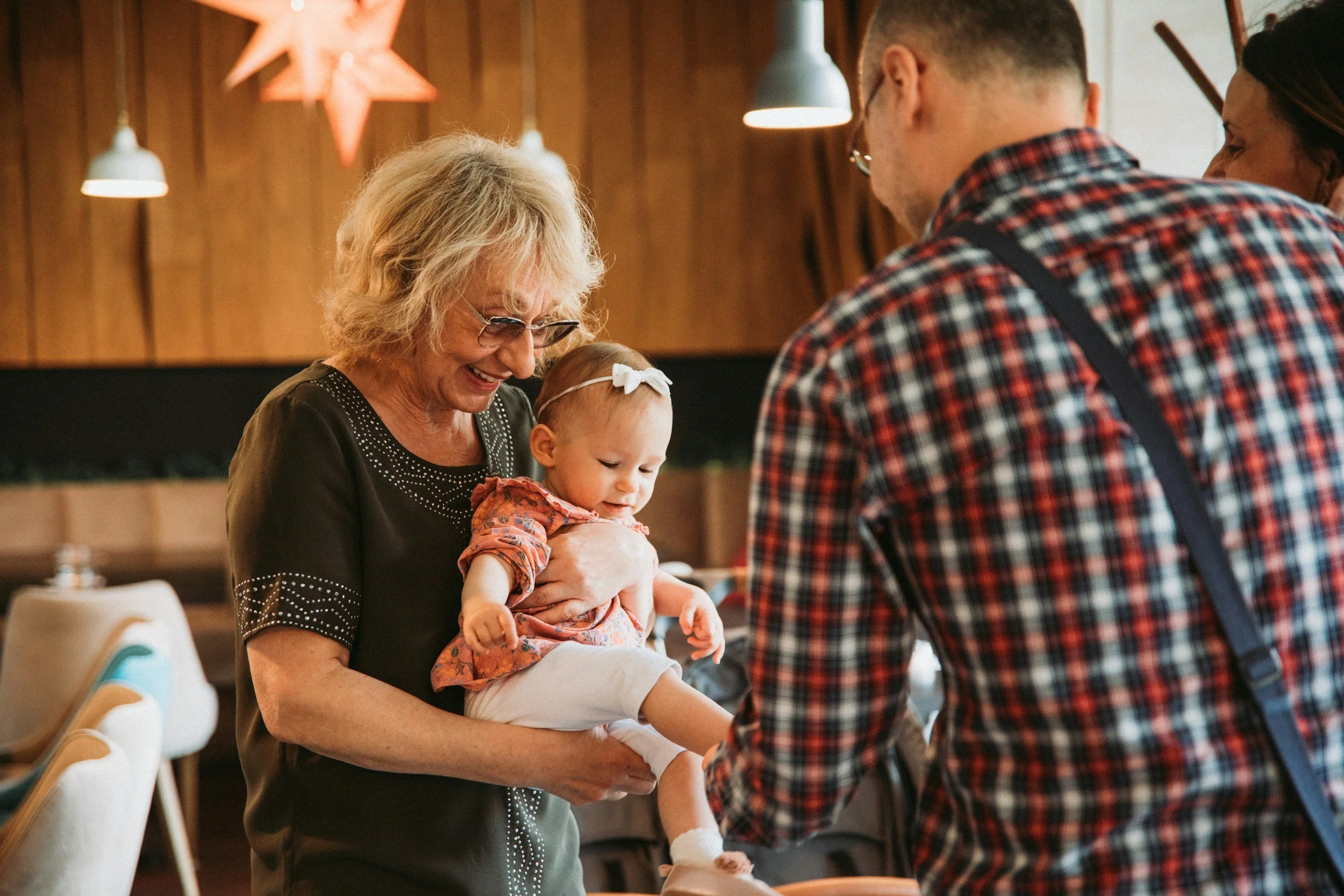 An elderly woman holding a baby girl, with a man and another woman in the background, in a cozy indoor setting decorated with stars hanging from the ceiling.