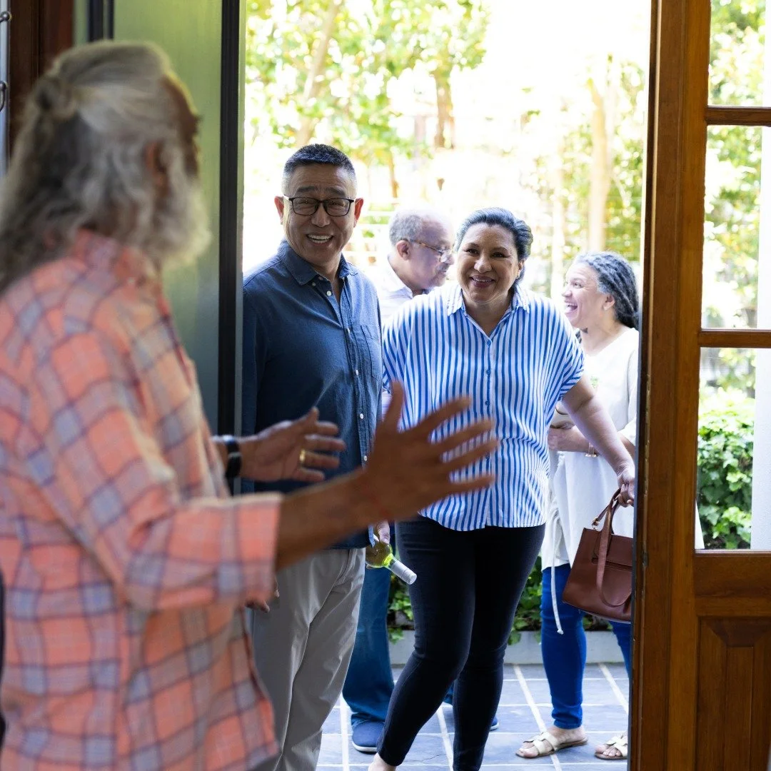 A group of people greet each other outside a house, smiling and chatting on the porch with lush green trees in the background.