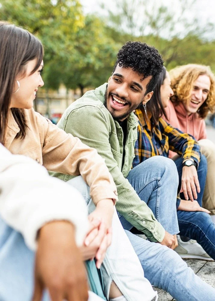Group of four young adults sitting outdoors on a bench, engaging in conversation and smiling, with trees in the background.