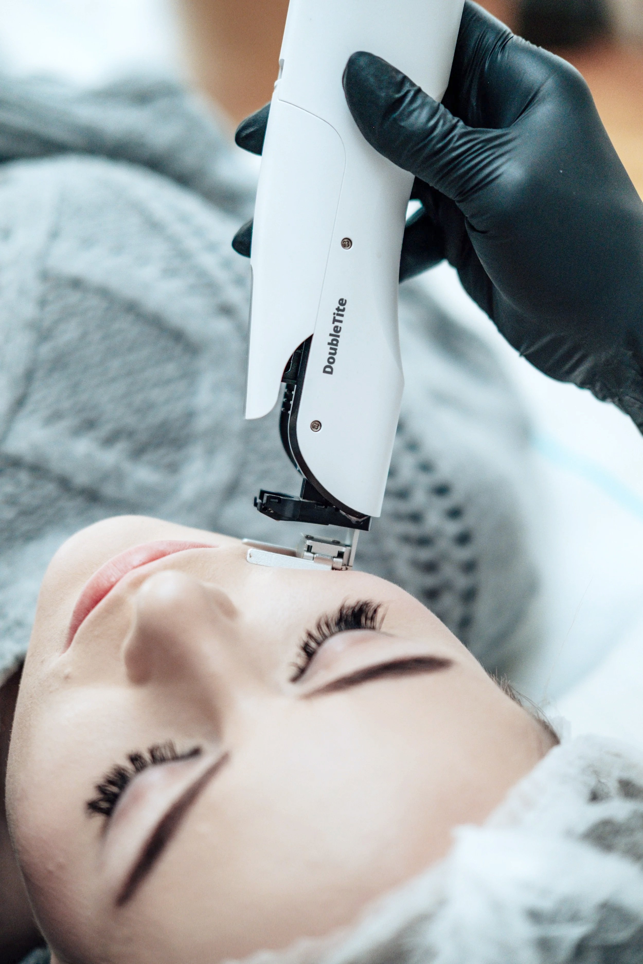 Eine Frau liegt mit geschlossenen Augen, während ein Arzt mit einer schwarzen Handschuhen eine Gesichtsbehandlung durchführt.