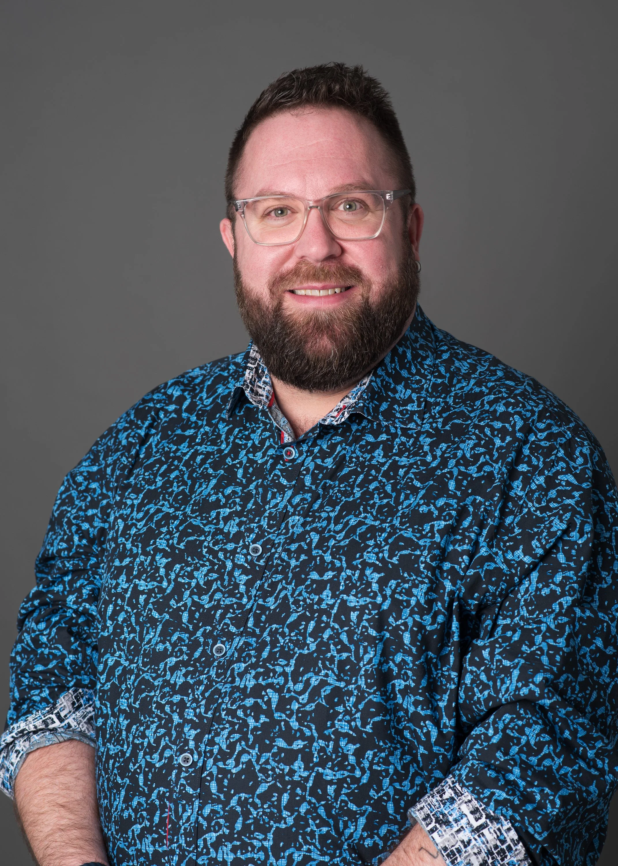 Person smiling at the camera in a studio portrait, wearing glasses and a blue patterned button-up shirt, standing against a neutral grey background.