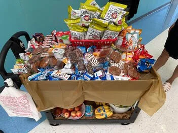 A shopping cart filled with various packaged food items, including snacks and candies, placed on a brown paper covering with some apples and a package of bananas at the bottom.