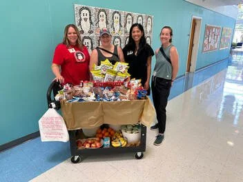 Four women standing behind a table filled with snacks and cookies in a school hallway
