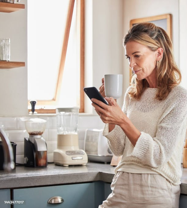 A woman with shoulder-length hair, wearing a beige sweater, is standing in a kitchen, looking at her phone, and holding a white coffee mug.