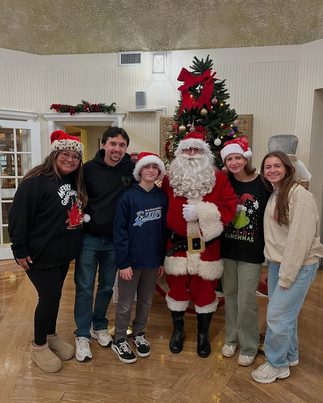 Group of six people, including Santa Claus, posing in front of a decorated Christmas tree, all wearing festive holiday clothing and Santa hats.