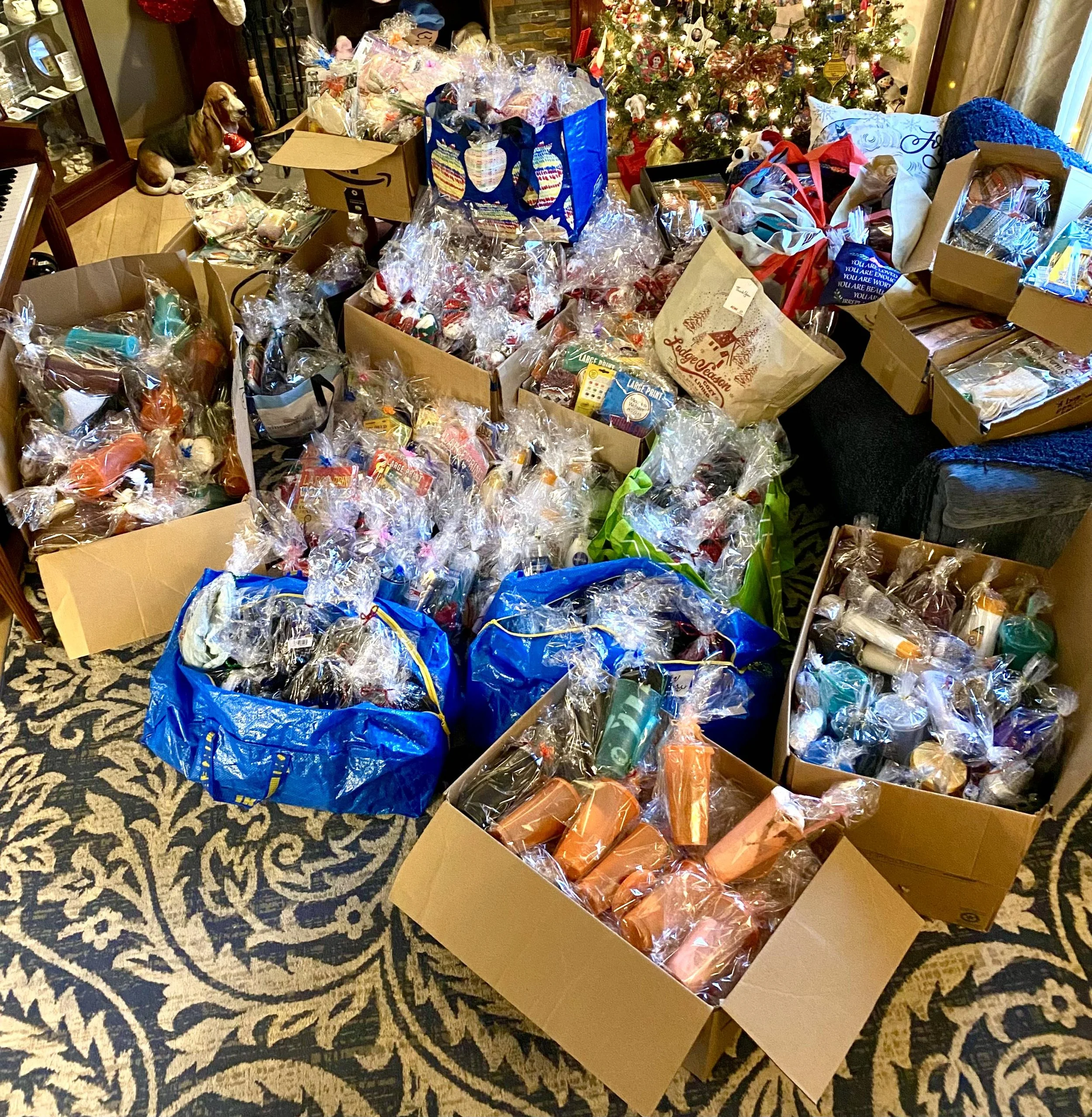 Multiple boxes and bags filled with wrapped holiday gifts, a decorated Christmas tree, and a dog sitting on the floor among the gifts.