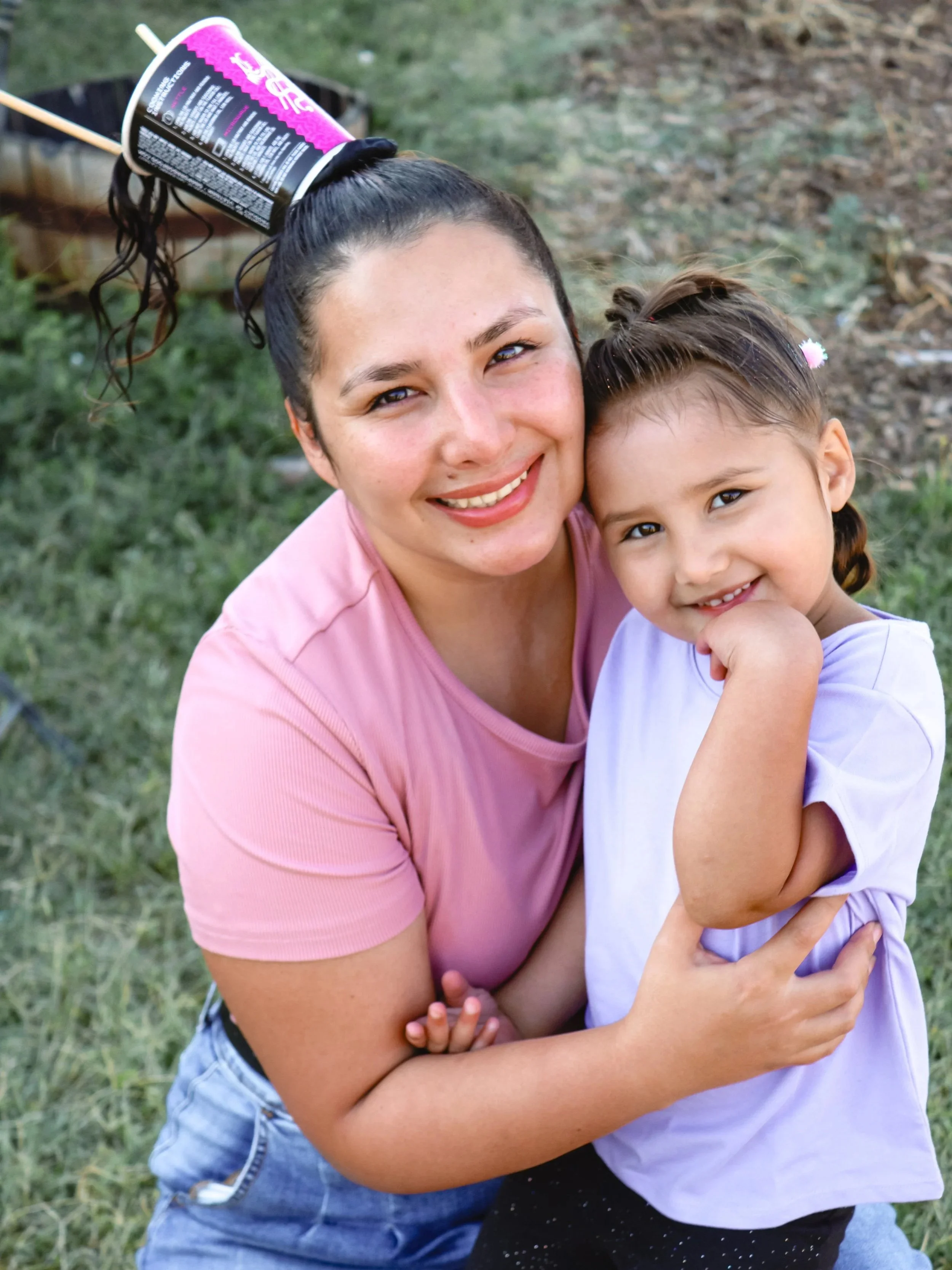 A woman and a young girl hugging each other outdoors, smiling at the camera. The woman has dark hair tied up with a cup on her head, and the girl has short hair with a small flower clip.