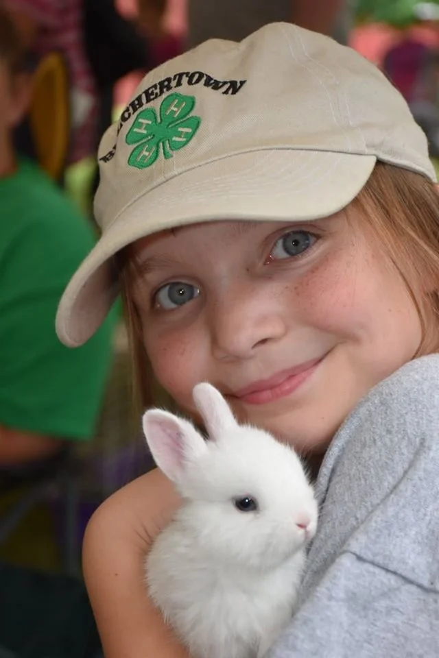 A young girl with blue eyes and freckles, wearing a beige cap with green and black embroidery, holding a small white rabbit.