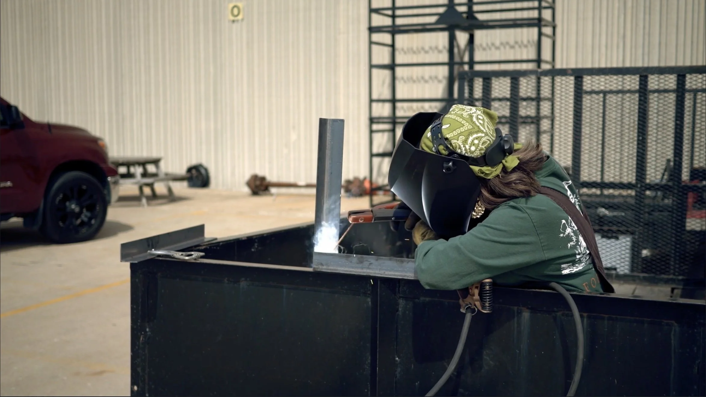 A person welding inside a large black container in a workshop. They are wearing a welding helmet, green bandana, green long sleeve shirt, and gloves.