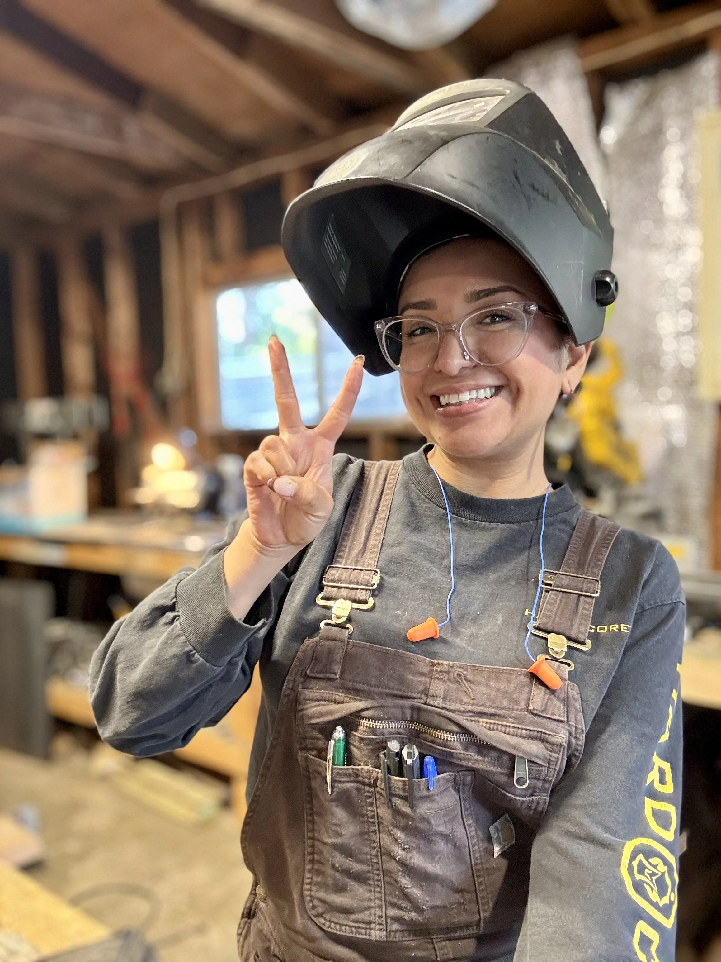 Jessica Rodriguez Hernandez. Dos Locos. Dos Locos Fabrication. Working in a workshop wearing a welding helmet raised above her head, smiling, making a peace sign, with pens in her pocket, and wearing ear protection around her neck.