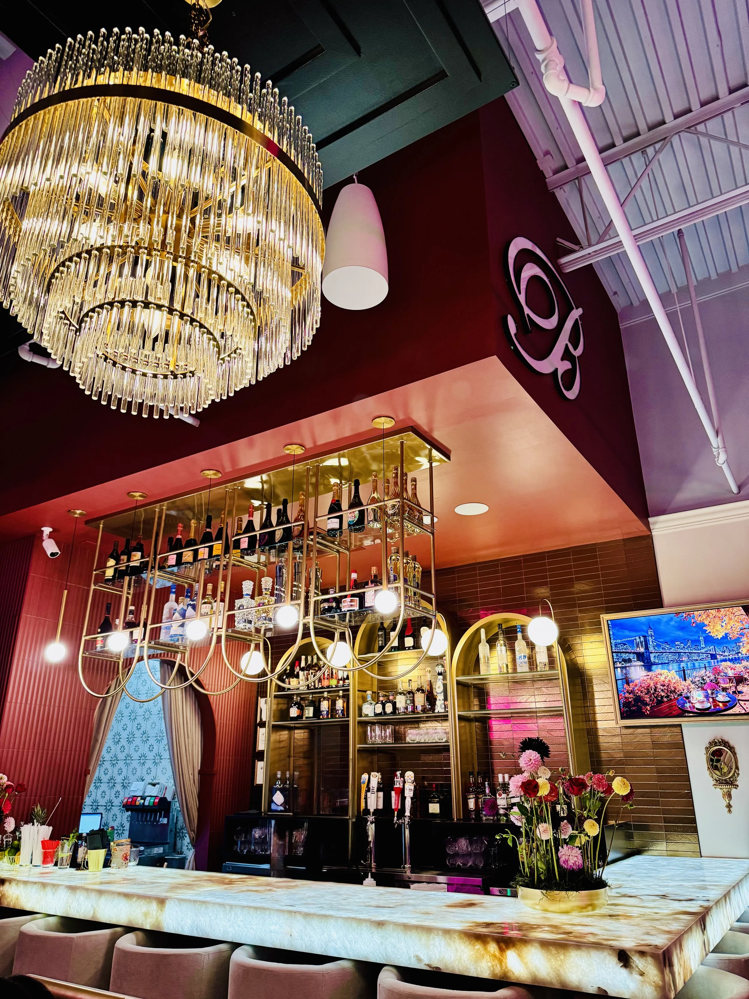 Elegant bar interior with a large crystal chandelier, gold and black shelves with liquor bottles, flowers on the counter, and a flat-screen TV displaying a scenic harbor view. Restaurant Shelving. Custom Bar Shelves. 