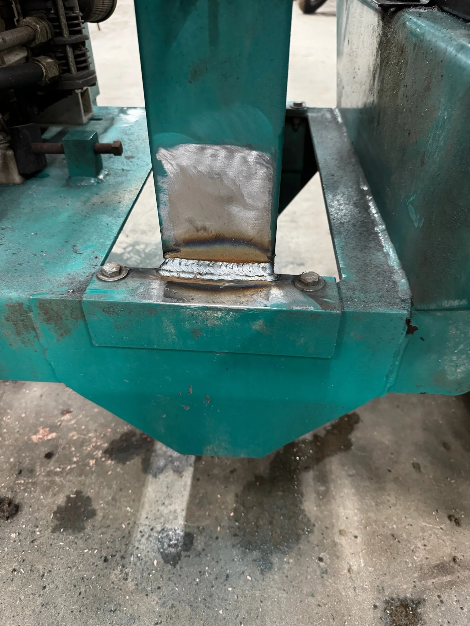 Close-up of a welded metal joint on a blue-green industrial machine or structure, with visible welding bead and heat discoloration.