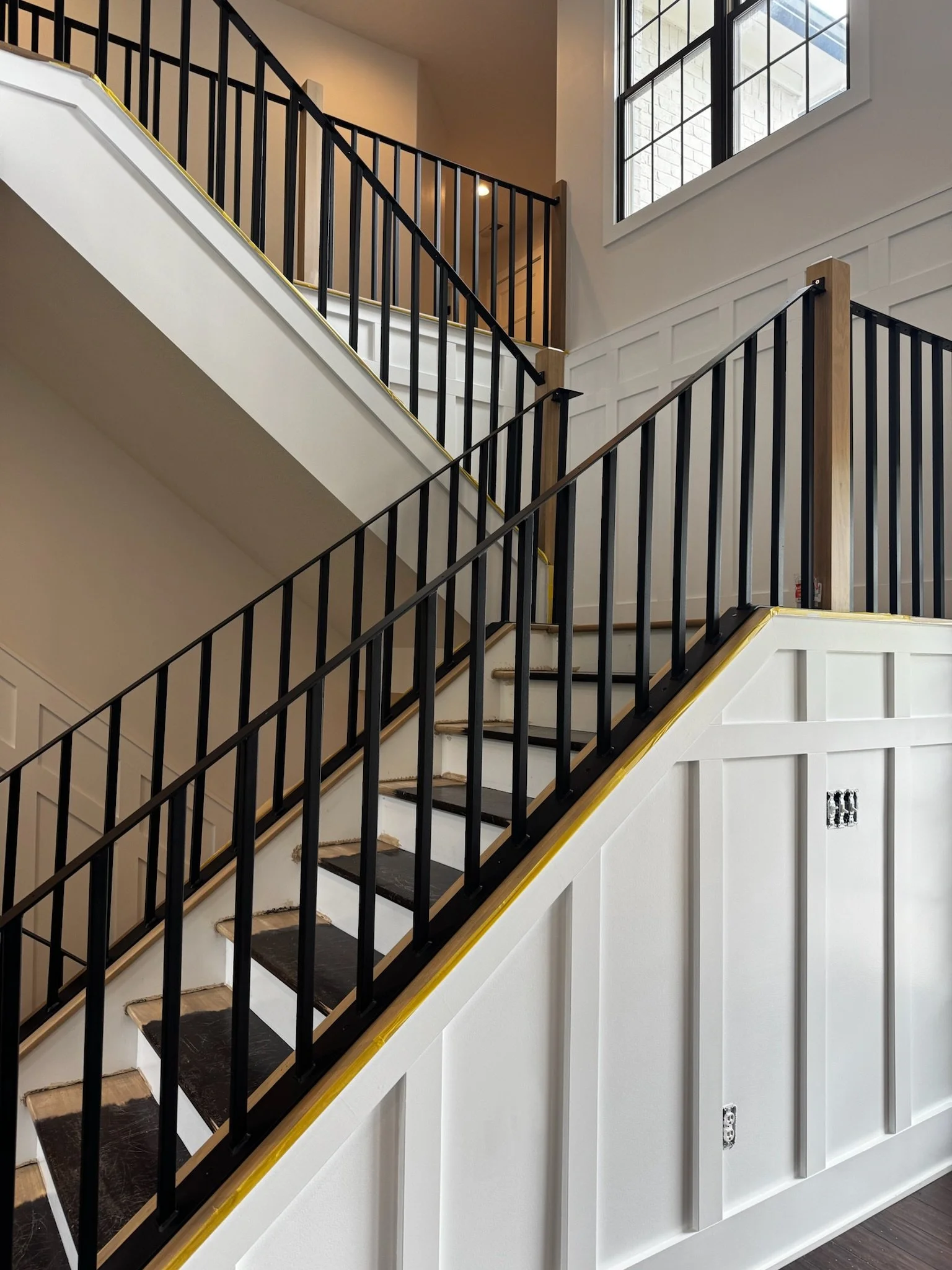 Interior of a multi-story house under construction showing a staircase with black railings, white wall paneling, and large windows at the top.