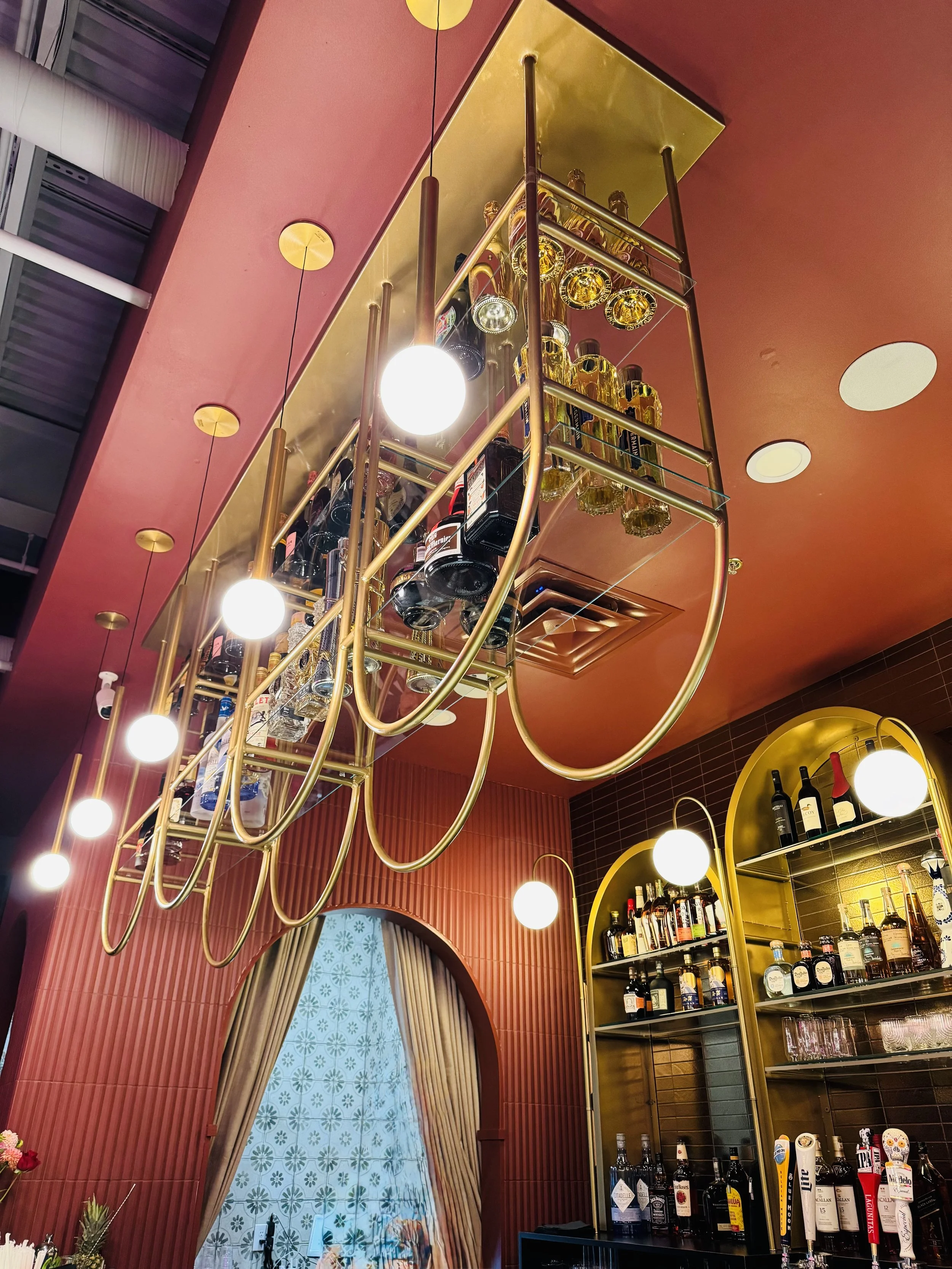 Overhead view of a bar with a gold and glass shelving unit holding various bottles of alcohol, illuminated by hanging spherical light bulbs, with a red and gold ceiling and a curtained window in the background.