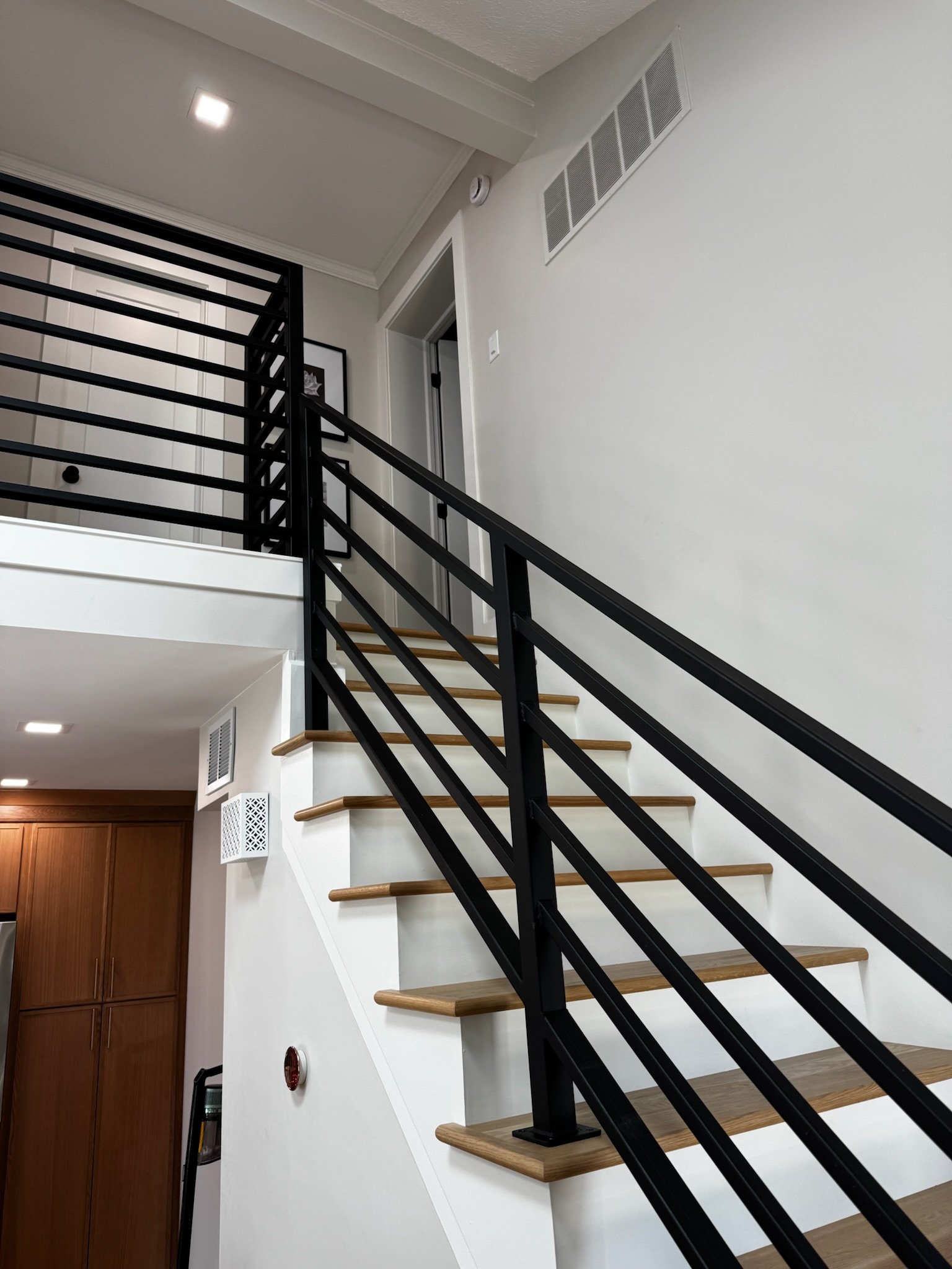 Interior view of a staircase with wooden steps and black metal railing, leading to an upper floor with a white wall, air vent, and a door.