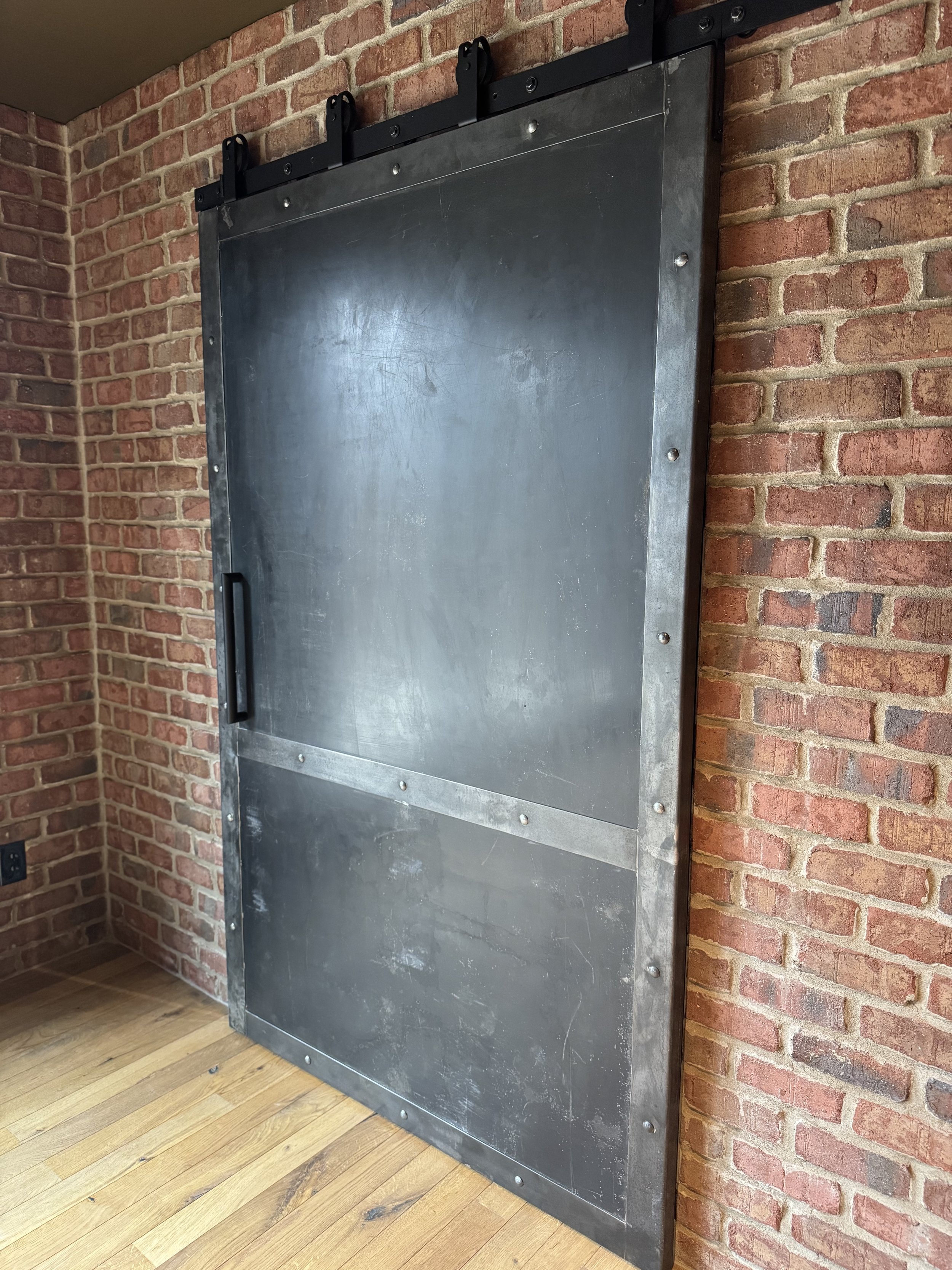 Metal sliding barn door installed on a brick wall with a black handle and black ceiling-mounted rollers.