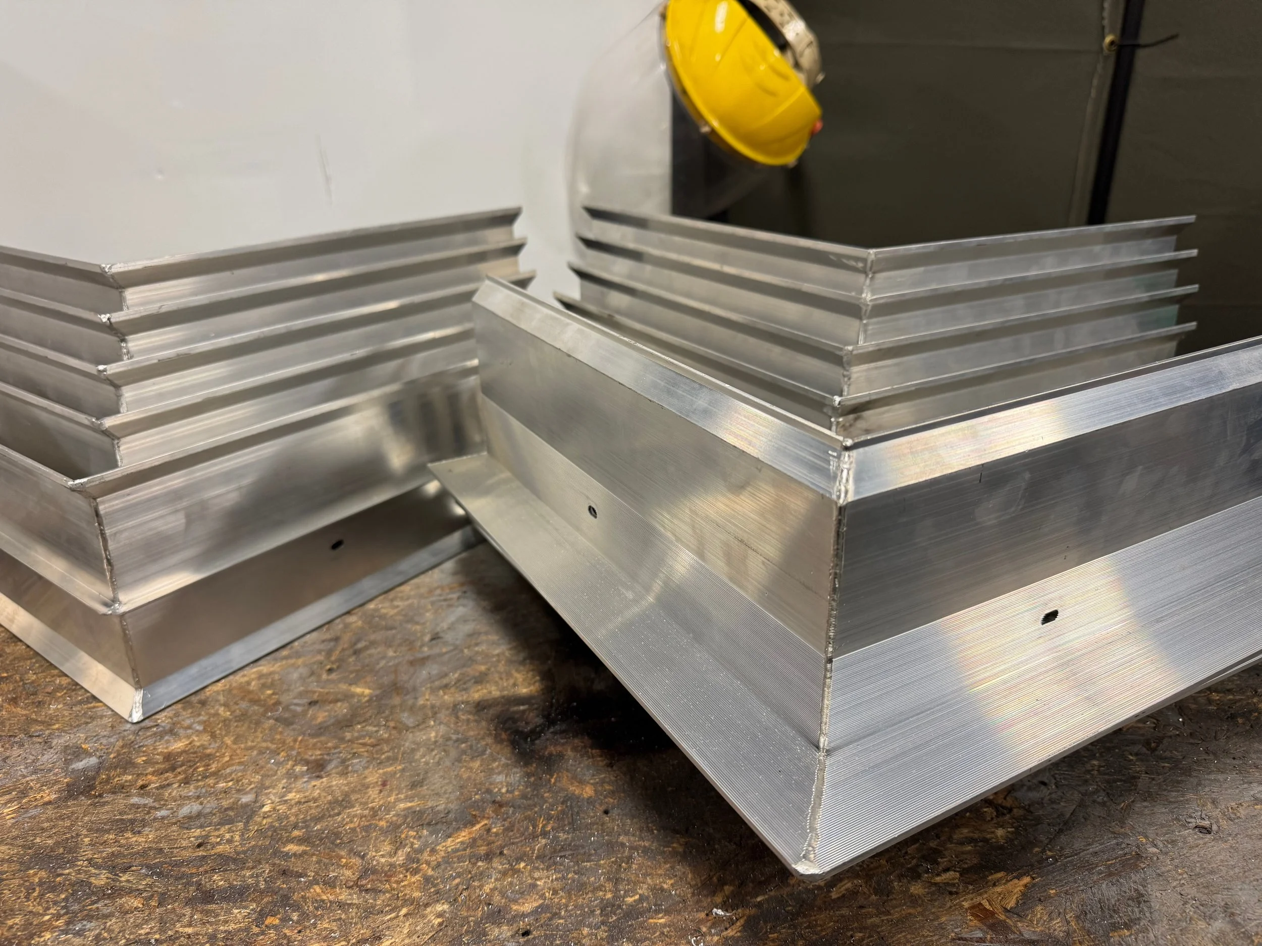 Two aluminum rectangular frames with multiple angled fins, placed on a rusty brown work surface. Roofing Miters. Welded Roofing Miters.