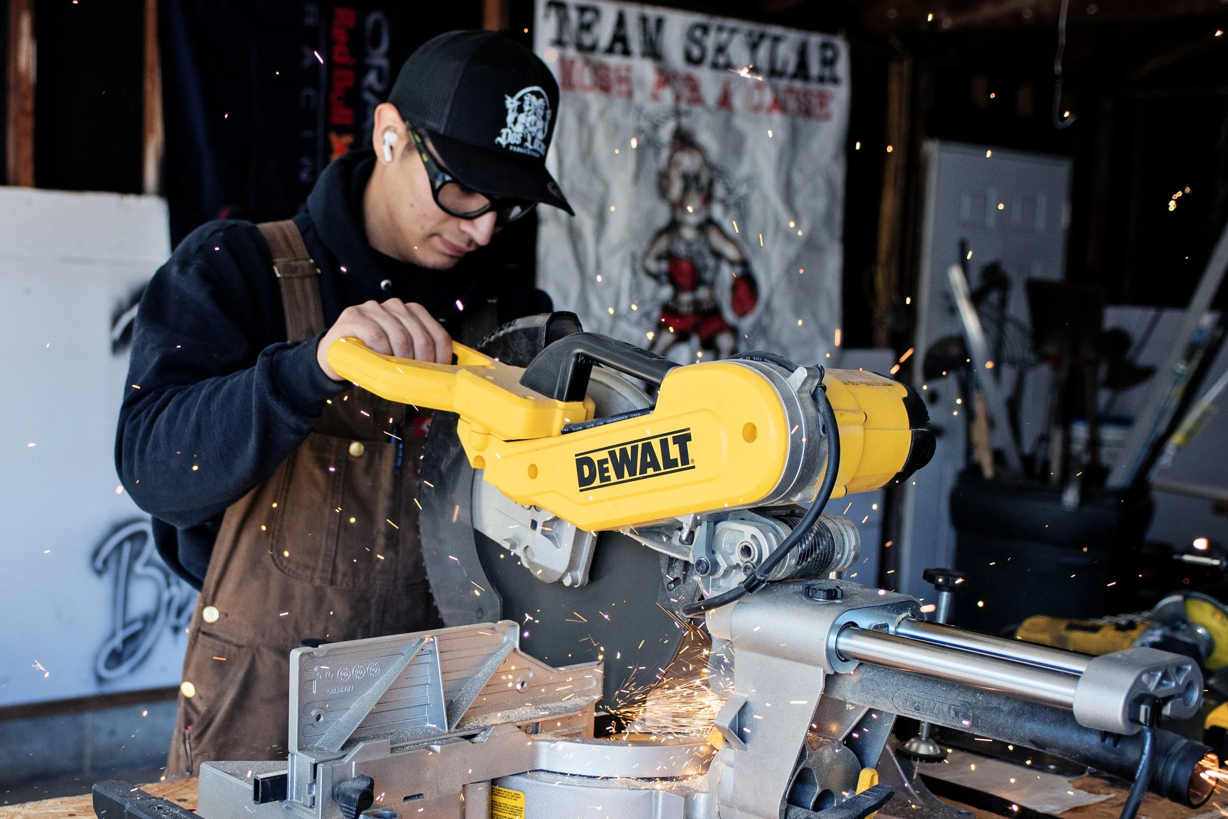 Alex Acosta. Dos Locos. Dos Locos Fabricationb.  Alex using a yellow DeWalt miter saw, cutting through wood with sparks flying, in a workshop with tools and a festive banner in the background.