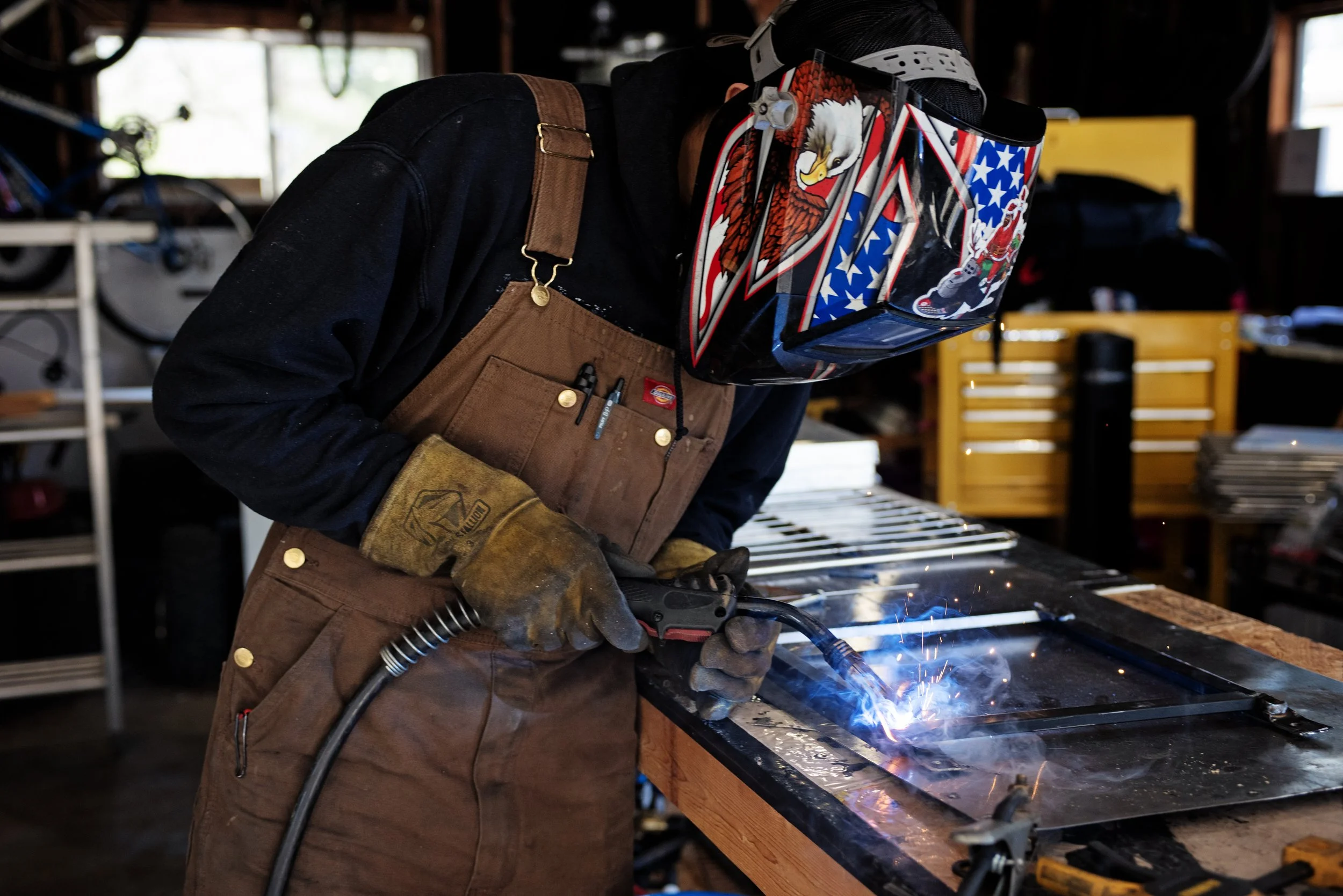 A person welding in a workshop, wearing a patriotic helmet with American flag and eagle designs, brown apron, and protective gloves, working on a metal frame at a wooden workbench.