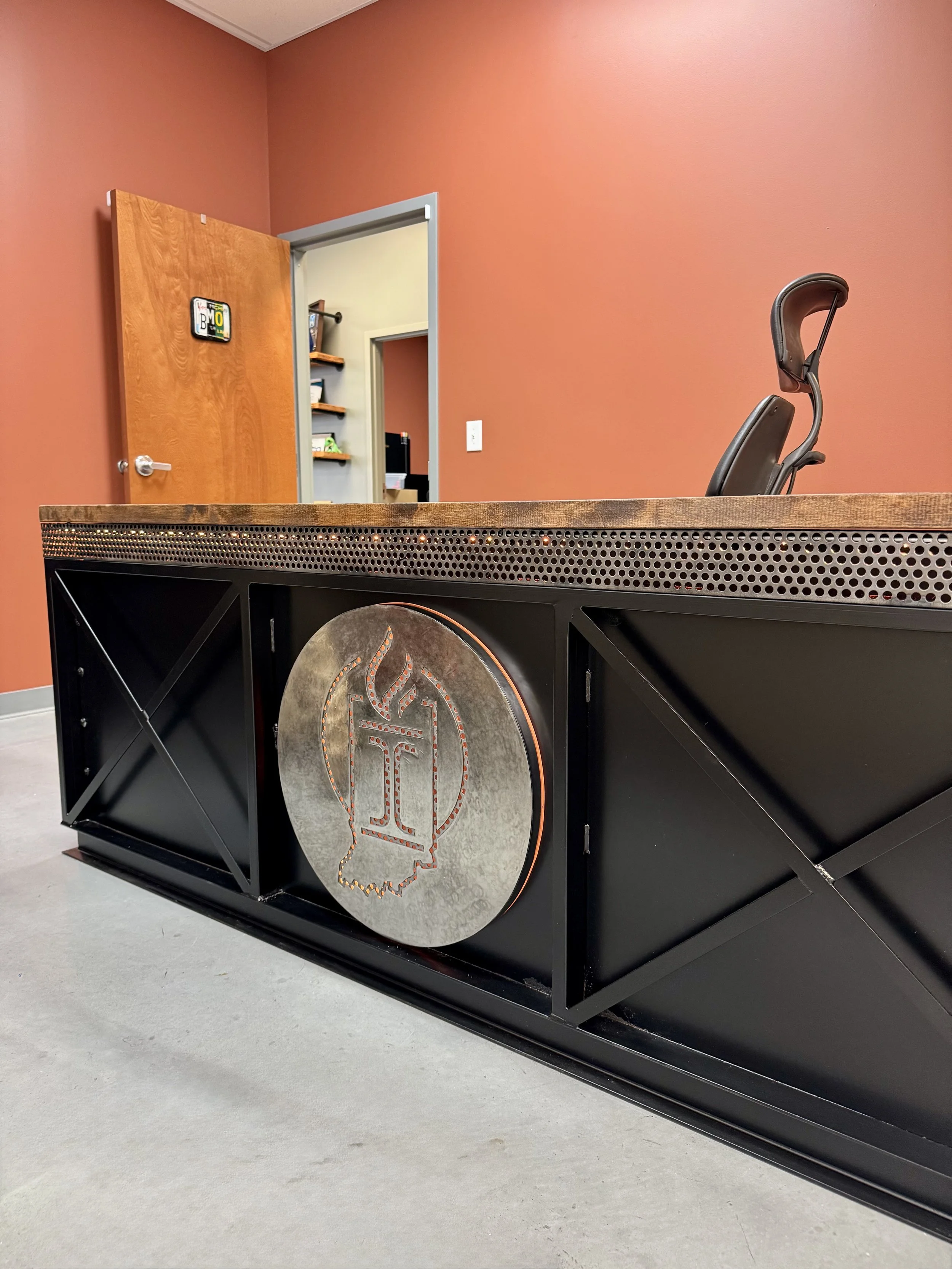 Modern office reception desk with a black metal frame and a large circular emblem with a torch and flame design. Behind the desk is a black ergonomic chair, and the background features a room with coral-colored walls, an open wooden door, and a doorway leading to another room with shelves.