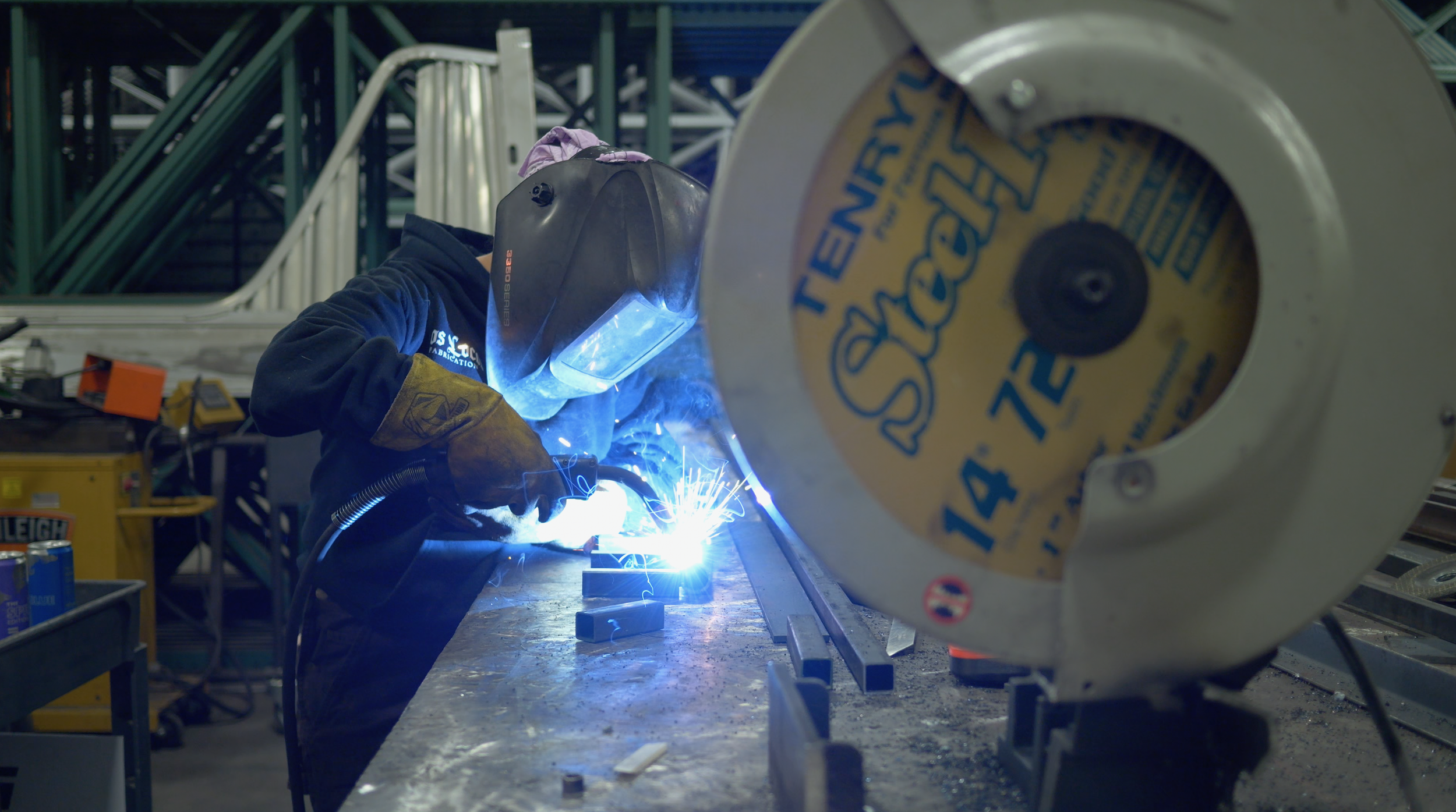 Worker welding metal in a workshop, wearing a welding helmet and gloves, with specialized equipment and metal supplies around.
