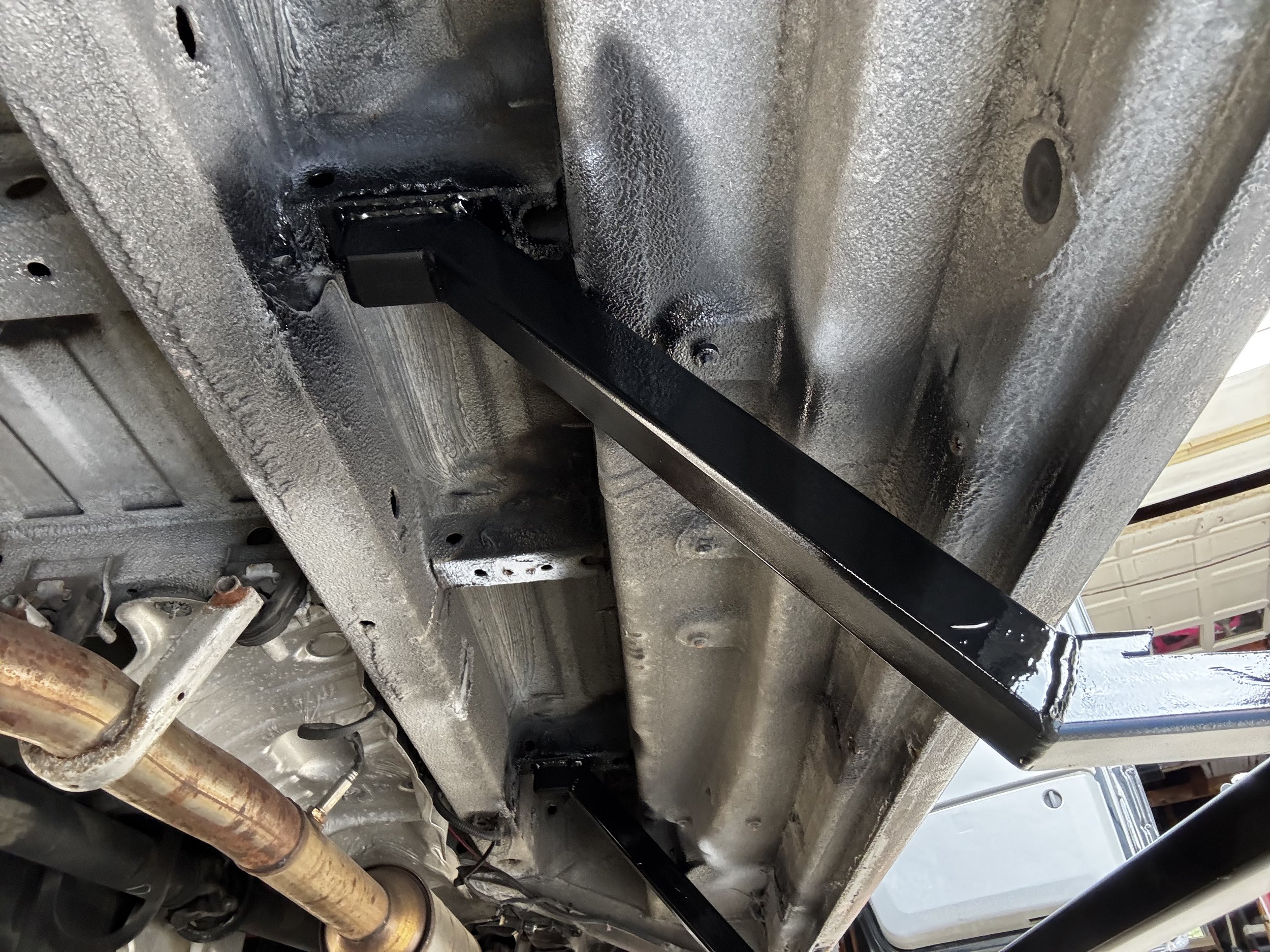 Underneath view of a vehicle's chassis showing a newly installed black metal support bar attached to the frame.