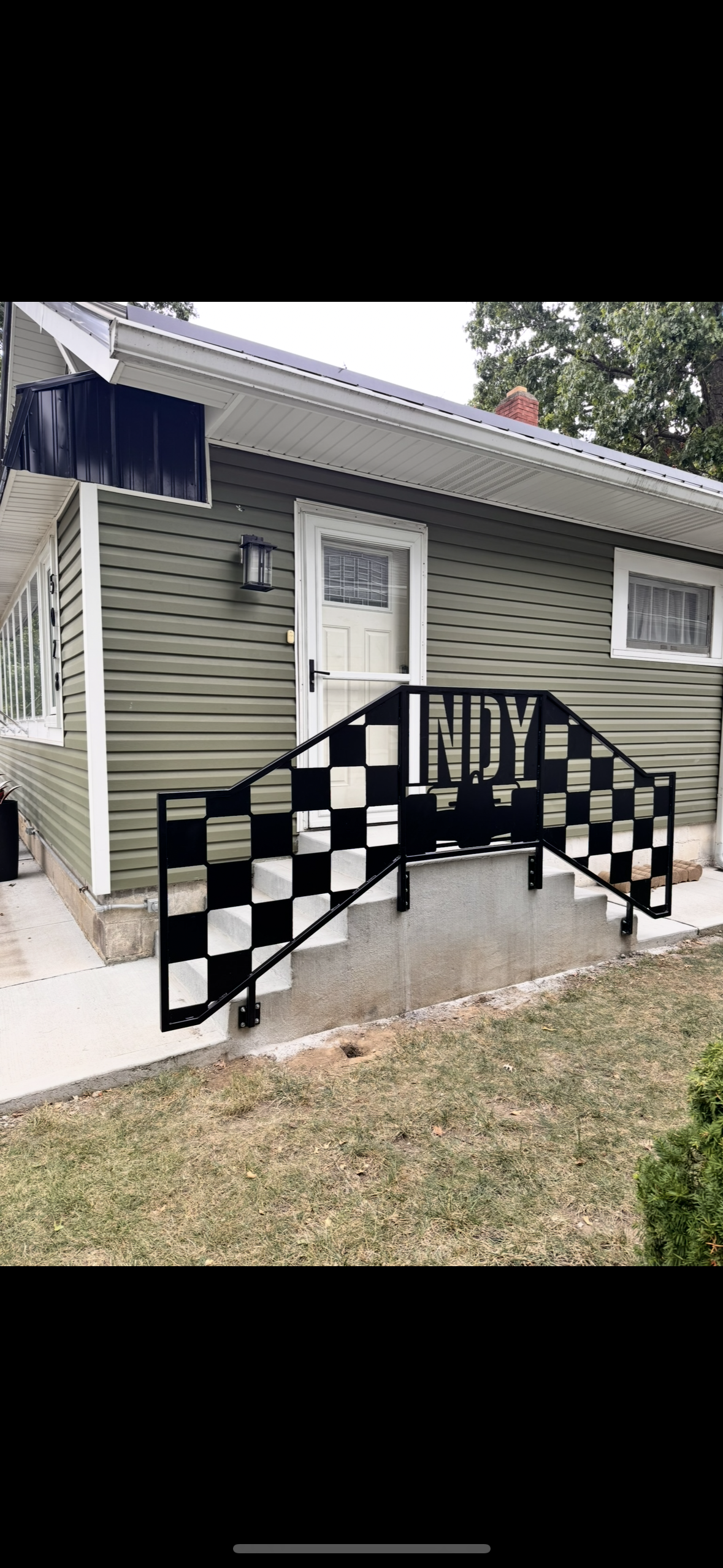 A house with green siding, white door, and small window, featuring a decorative black metal railing with a checkered pattern and the name 'INDY' on it, leading to the front steps.