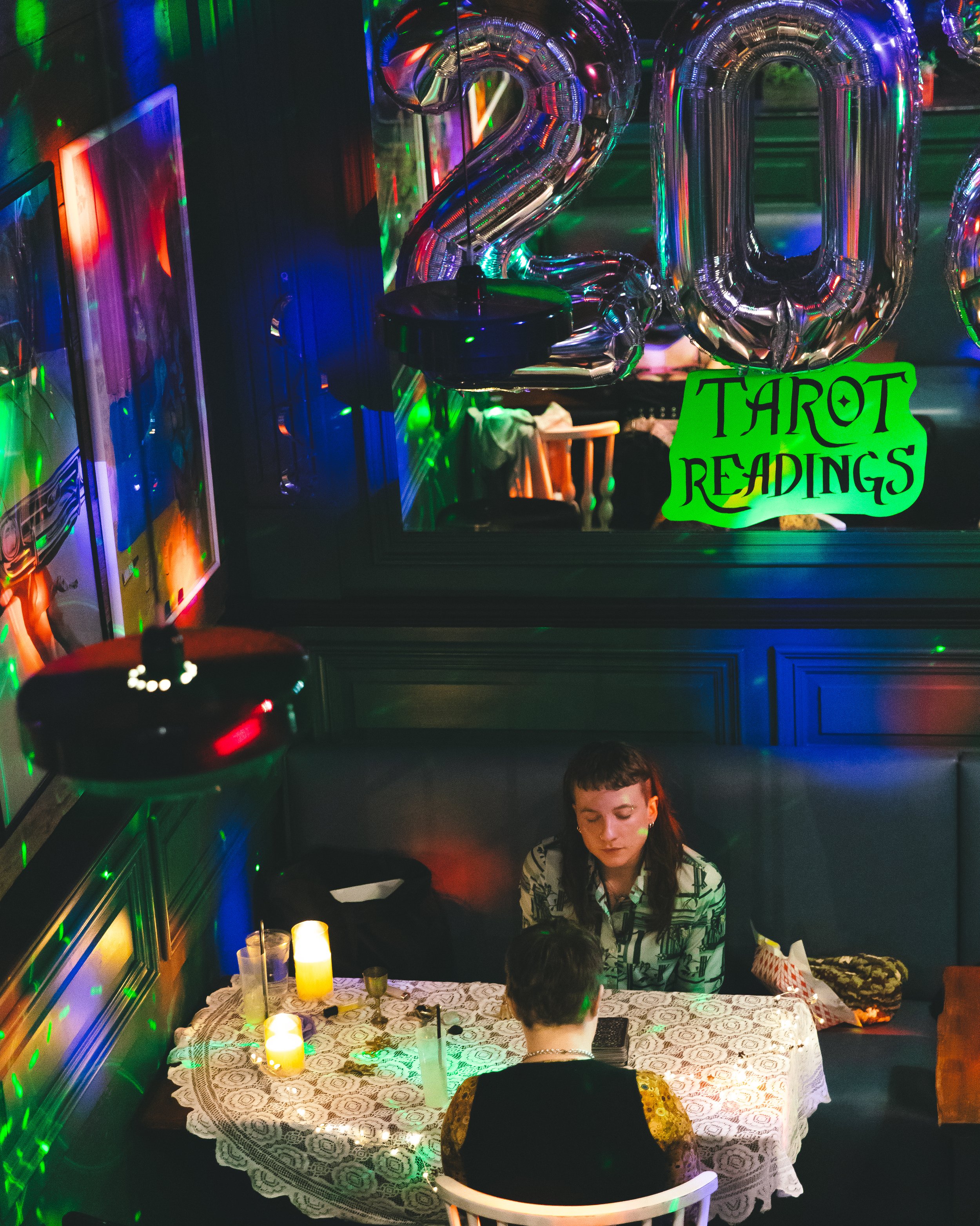 A table with two people reading tarot cards in a dimly lit room decorated for a 2020 celebration with large silver balloons. The table has a lace tablecloth, candles, and drinks. A neon sign on a mirror reads 'Tarot Readings.'