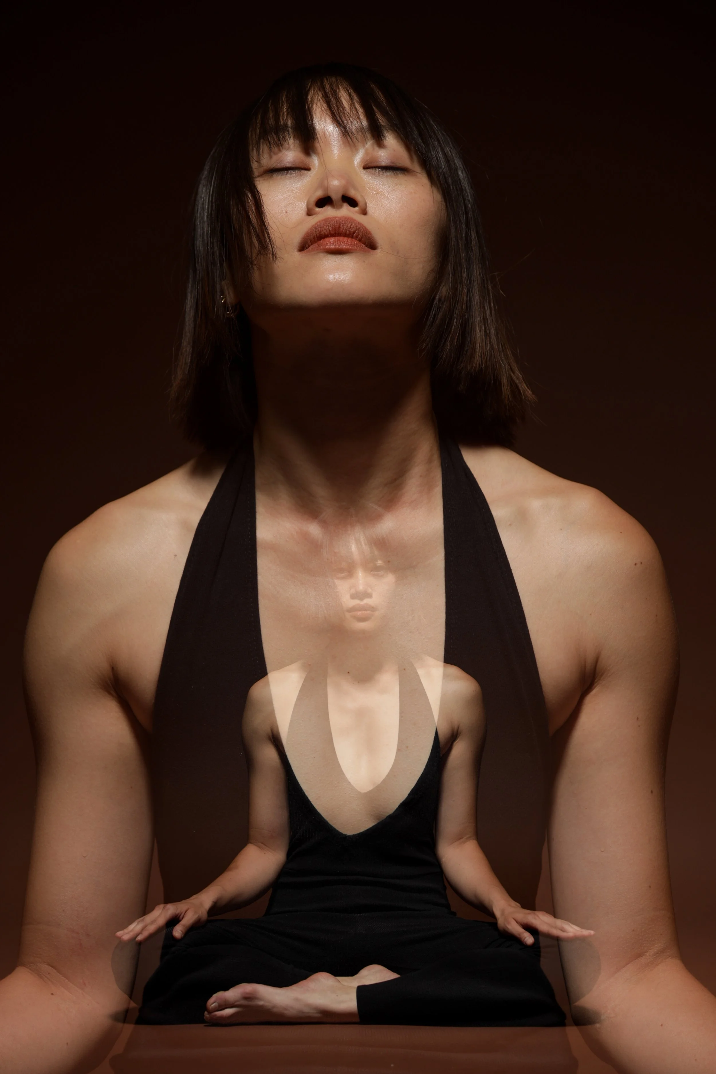 A woman with short dark hair and a black tank top sitting cross-legged, with a double exposure effect showing her meditation pose reflected within her body, against a dark background.