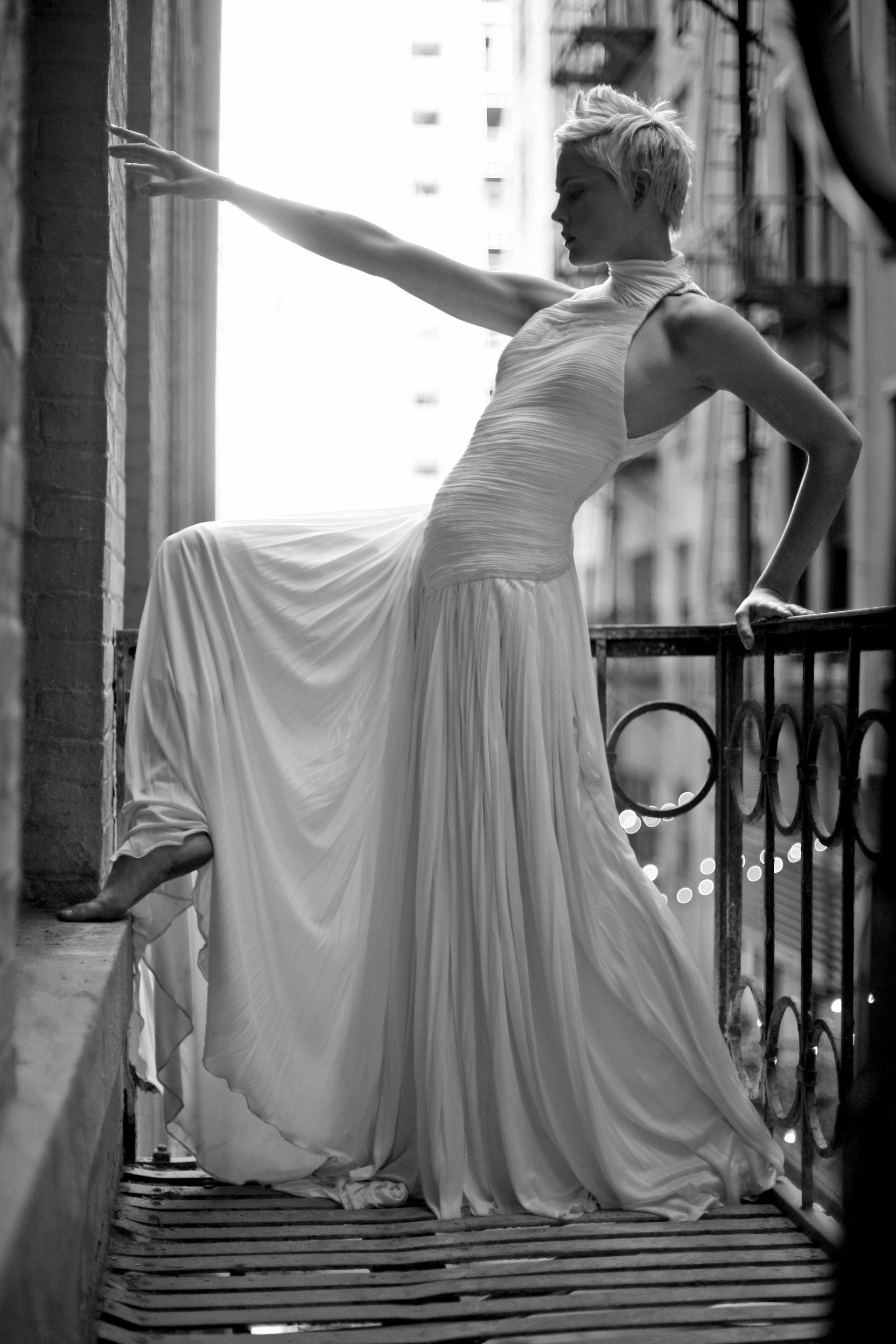 A woman with short blonde hair wearing a long, flowing white dress is standing on a fire escape, leaning on the railing with one hand and reaching out with the other. The photo is black and white, with city buildings in the background.