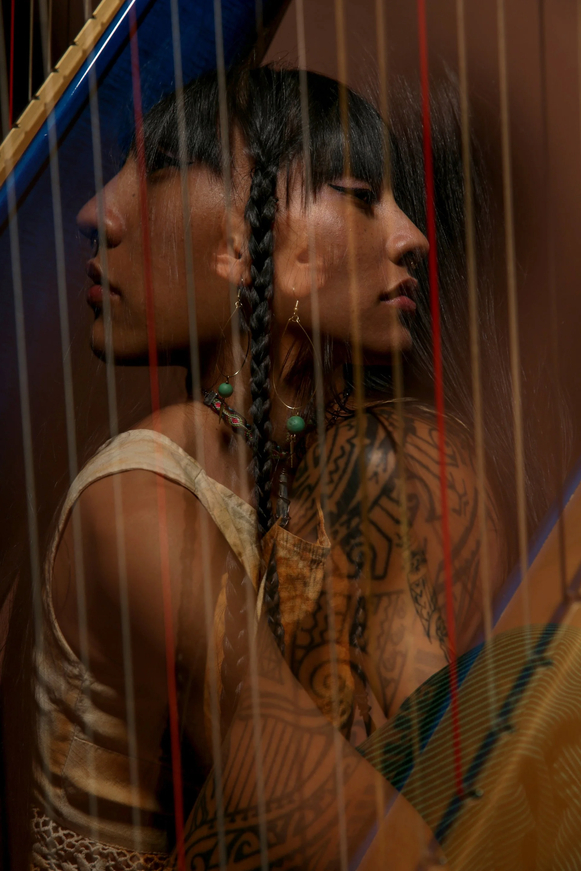 Two women with dark hair and tattoos behind a fence with vertical red and brown lines. One woman has short hair and the other has long hair with a braid. They are wearing earrings and looking to the right.