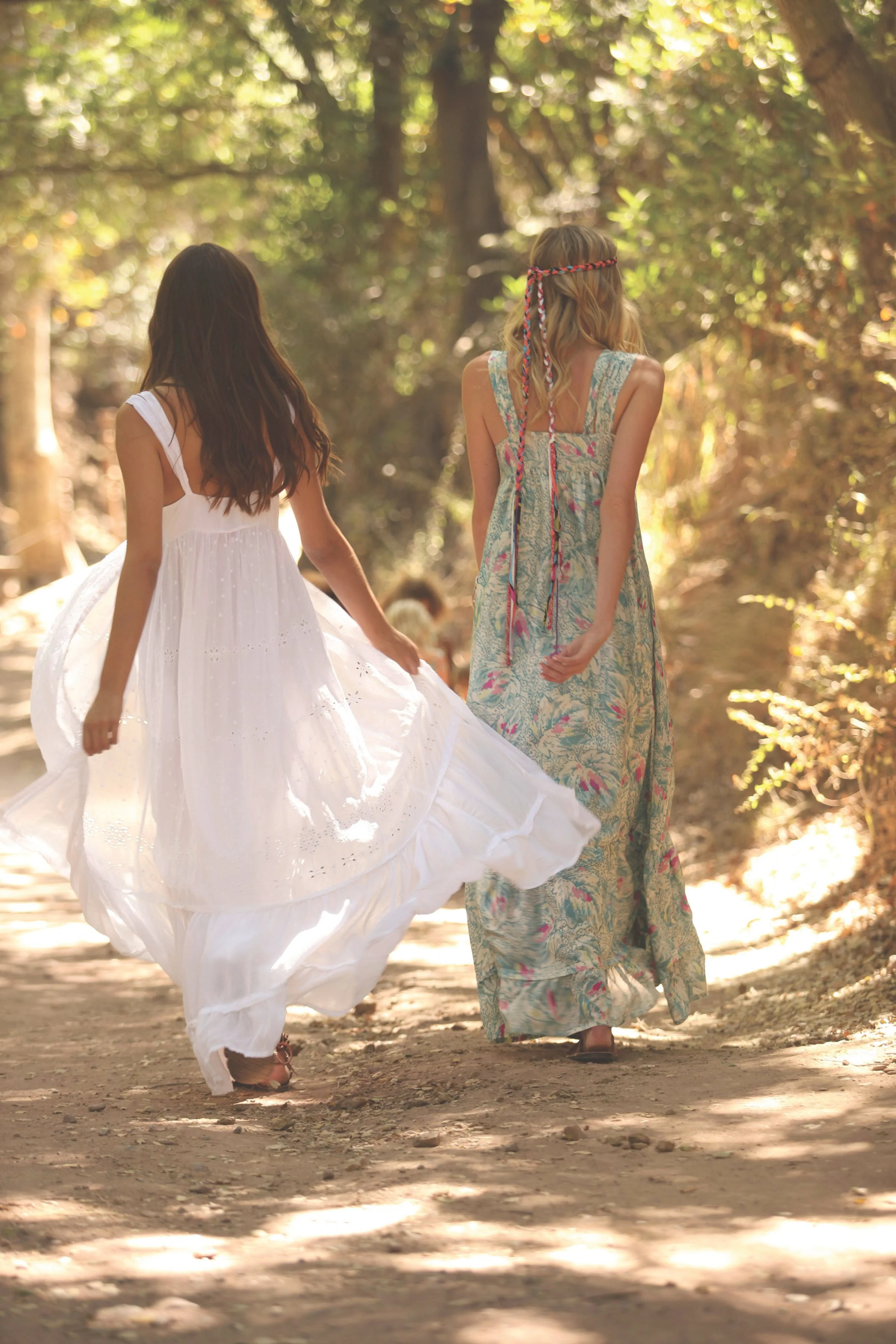 Two women walking along a wooded path, one in a flowing white dress and the other in a patterned maxi dress with a braided headband, surrounded by sunlight filtering through the trees.