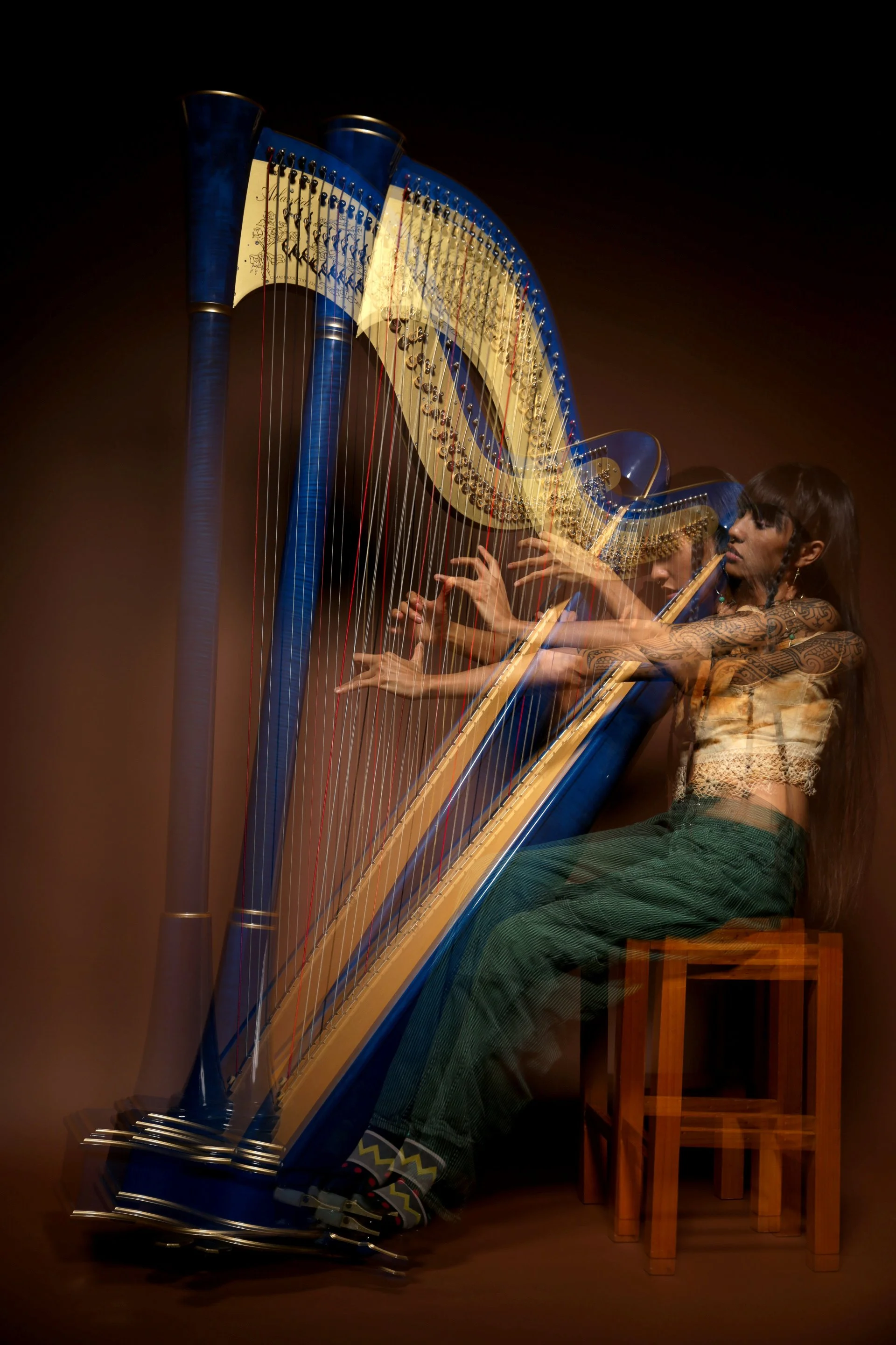 A woman with tattoos playing a large blue harp on a wooden stool.