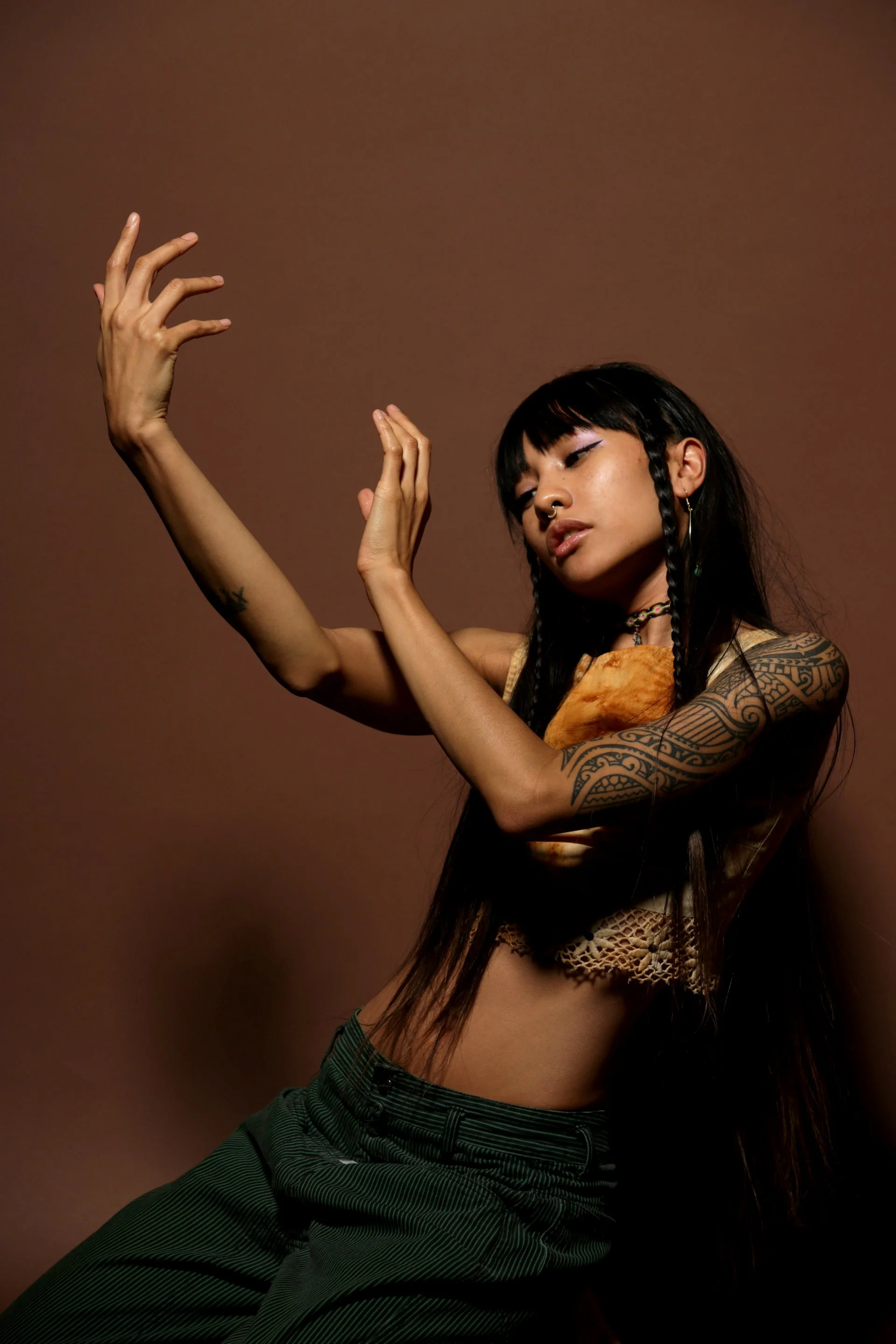 Young woman with black hair styled in braids, wearing jewelry, and dressed in a combination of clothing, posed with arms raised and hands moving delicately against a brown background.