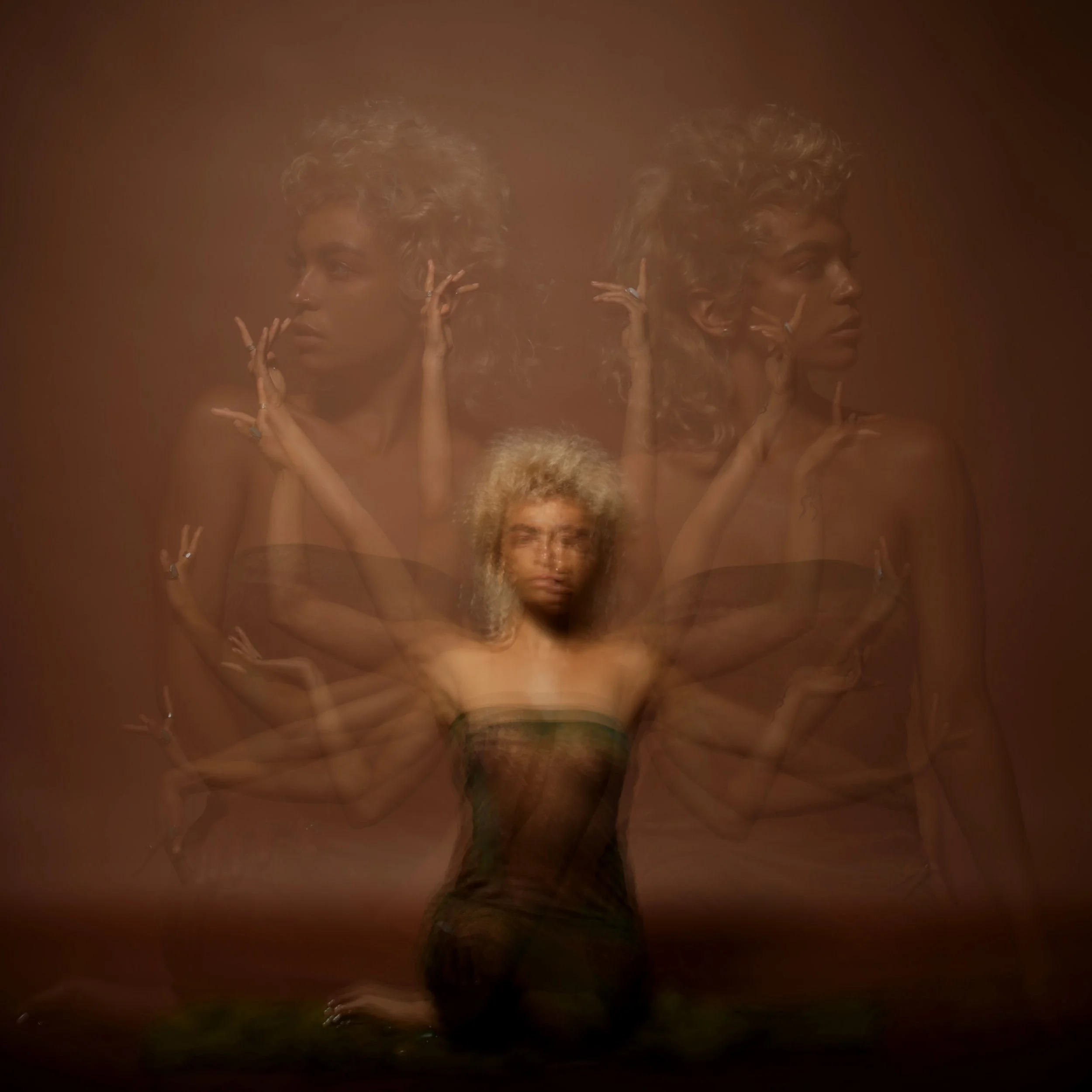 A woman with curly hair posing with her hands near her face against a warm, brown background, with a mirror reflecting her profile.