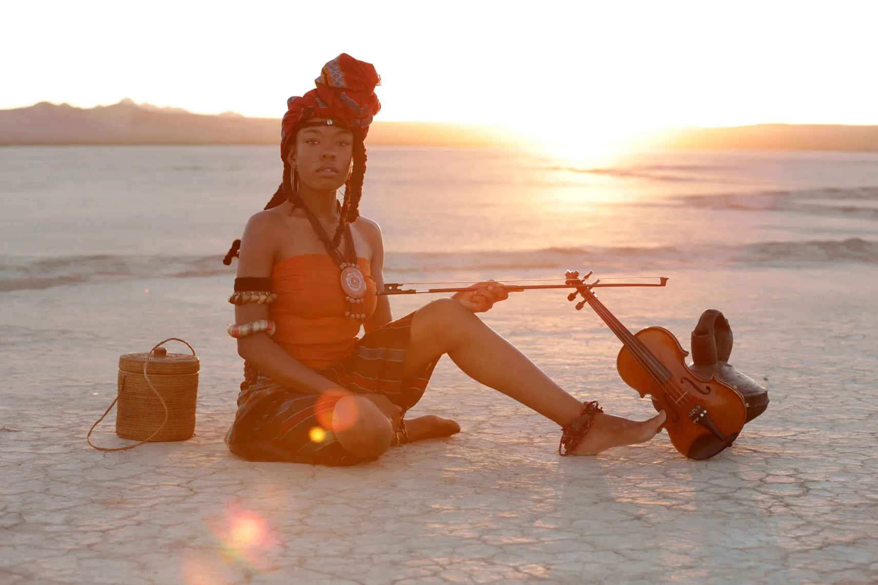 A woman sitting on a dry, cracked desert landscape at sunset, holding a violin and bow. She is wearing traditional accessories and an orange top. A woven basket is on her side, and a shoe is placed near her foot.
