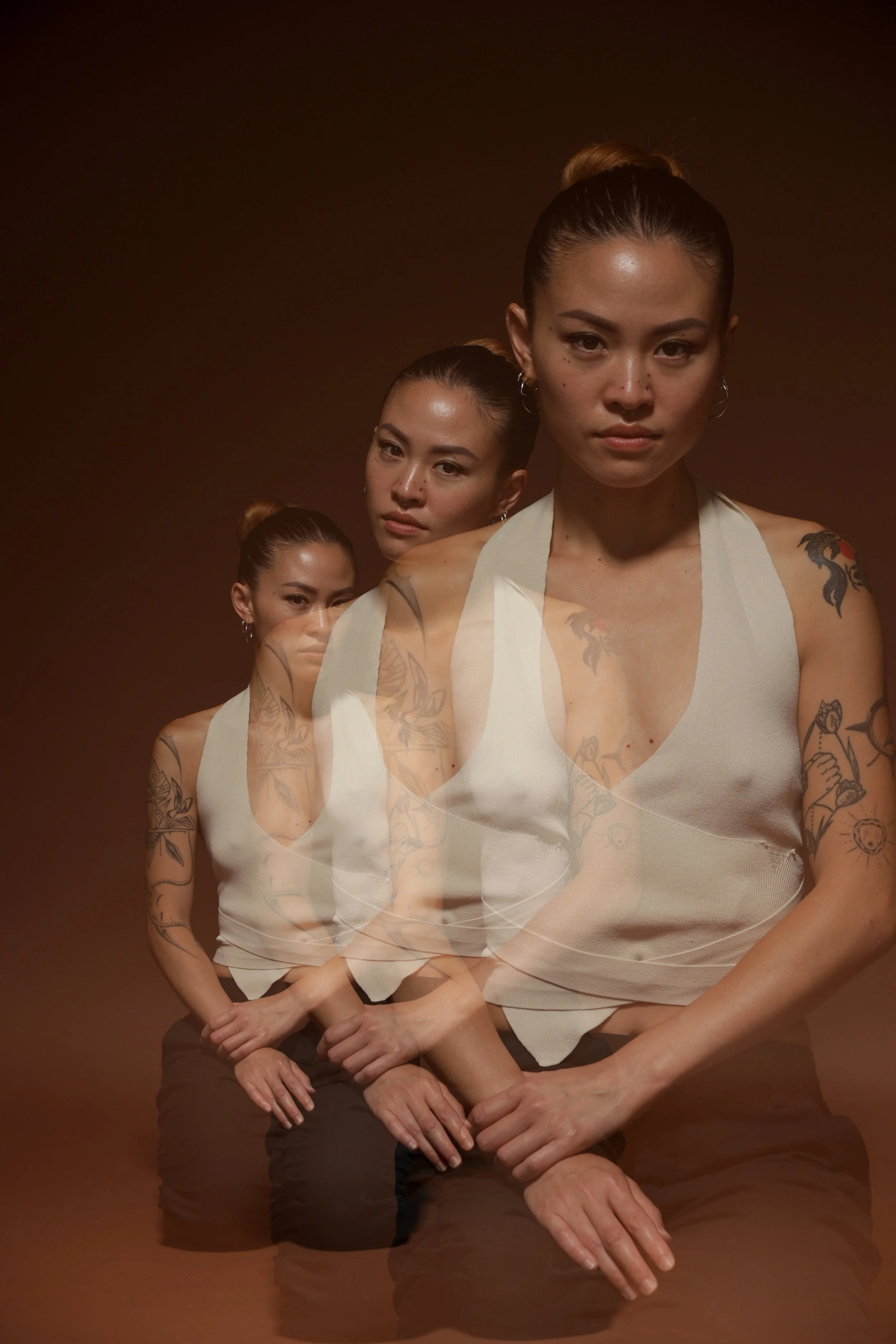 Multiple women with tattoos and similar clothing, sitting in a line against a dark brown background, with an overlapping, ghosted effect.