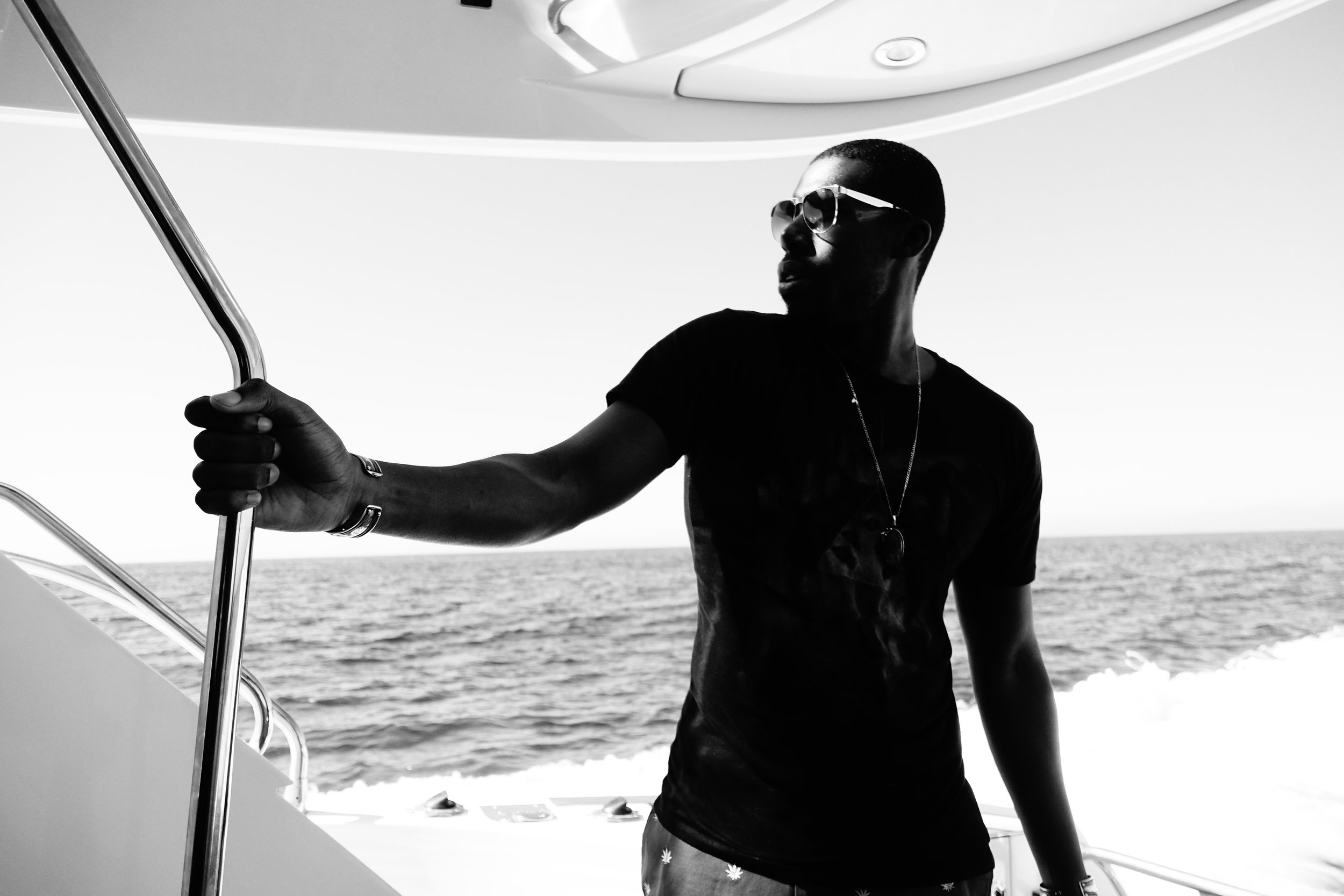 A man wearing sunglasses and a black t-shirt stands on a boat, holding onto a metal railing with a body of water and a distant horizon in the background.